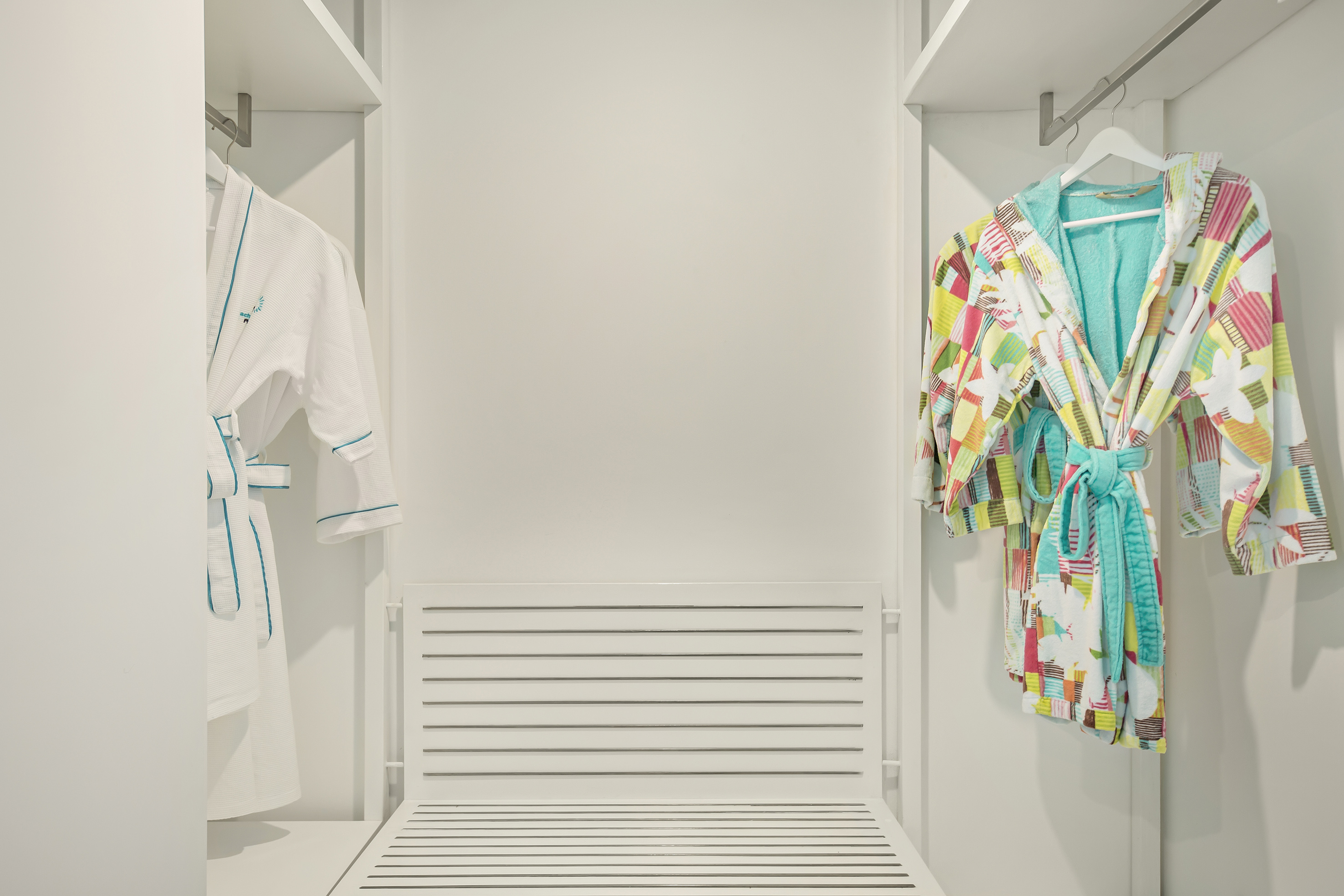 White and colorful bathrobes hanging in a hotel closet with a slatted bench.