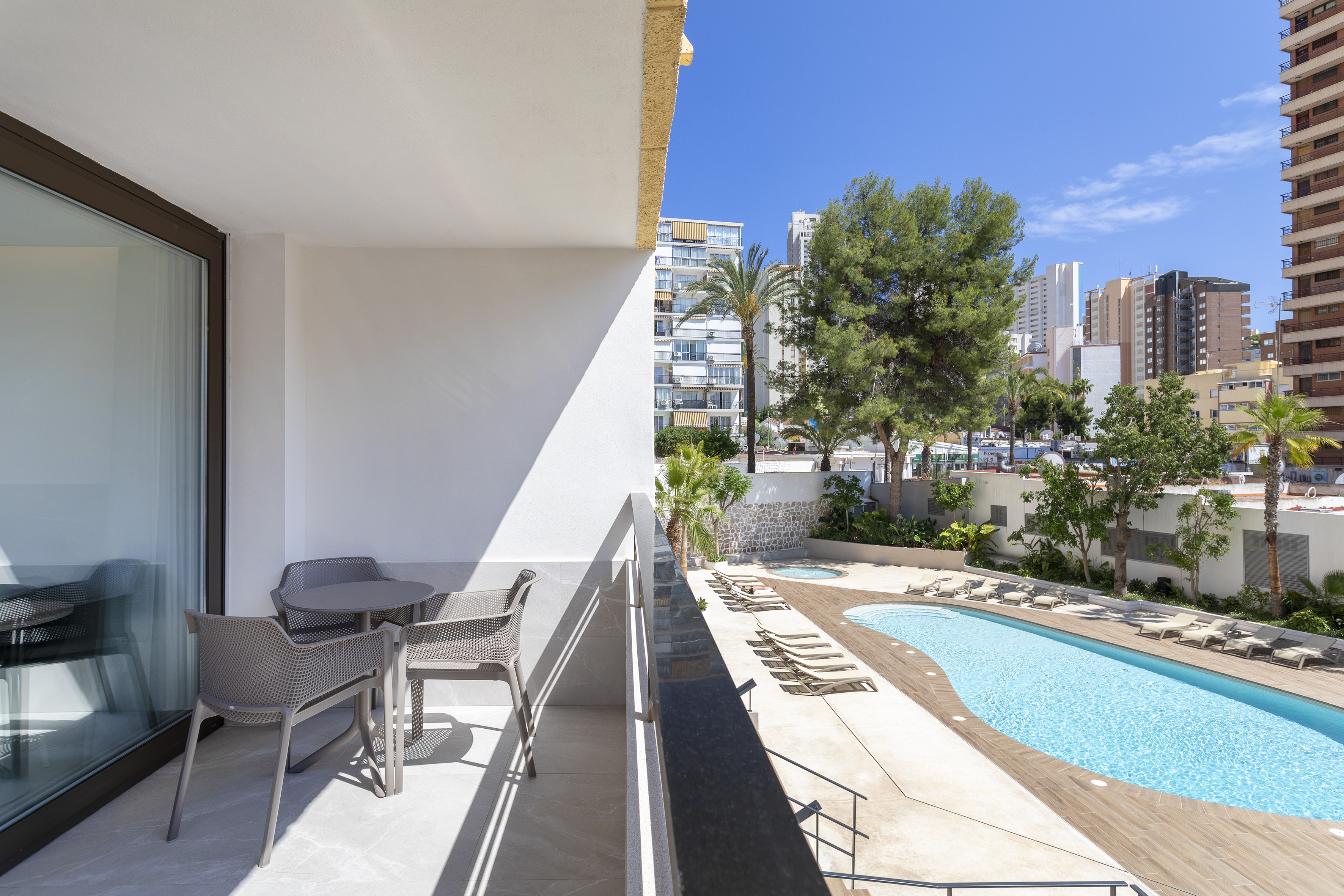 Sunny balcony with outdoor seating overlooking a hotel pool and city view.