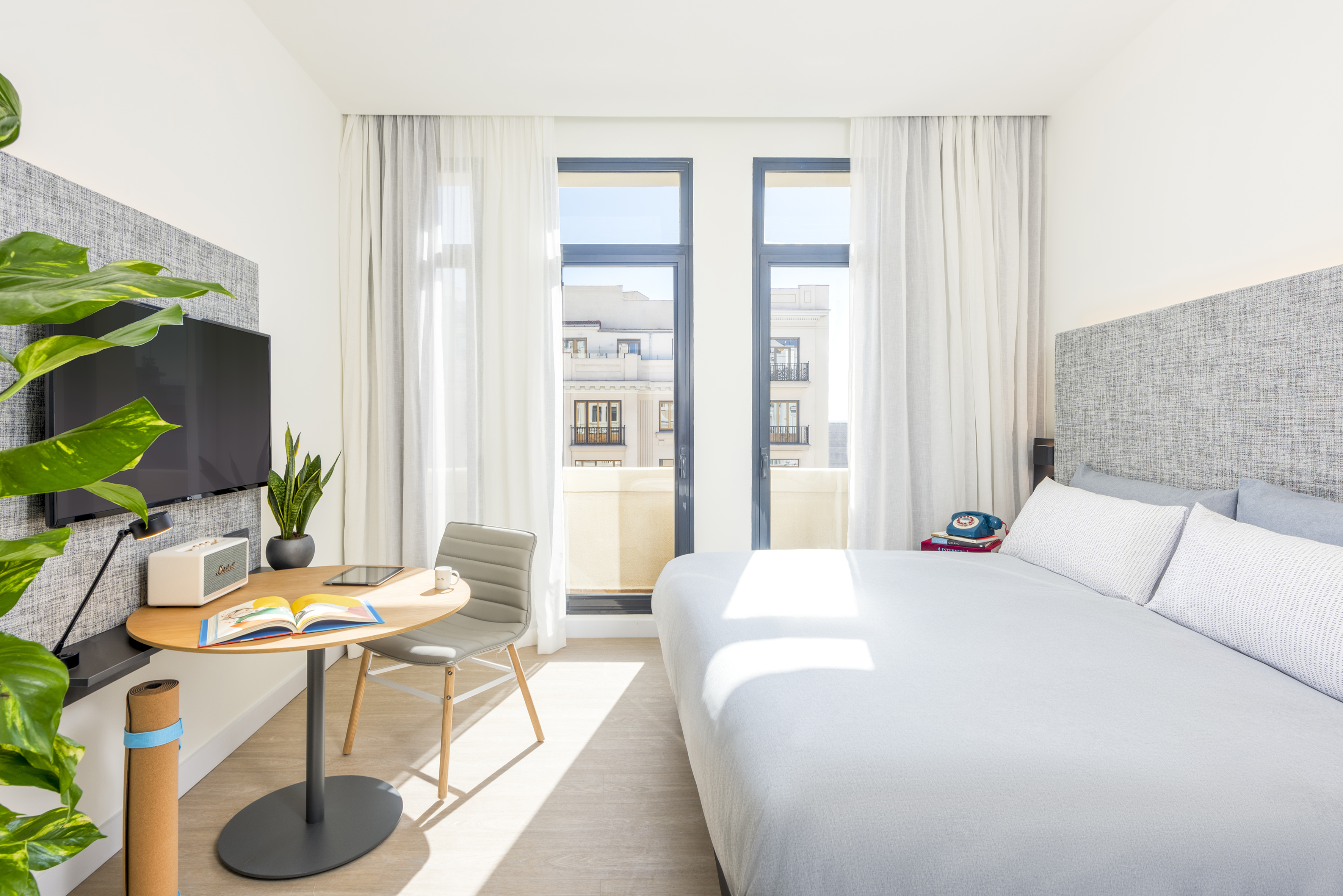 Bright modern hotel room with white bed, large window, desk, and wall TV.