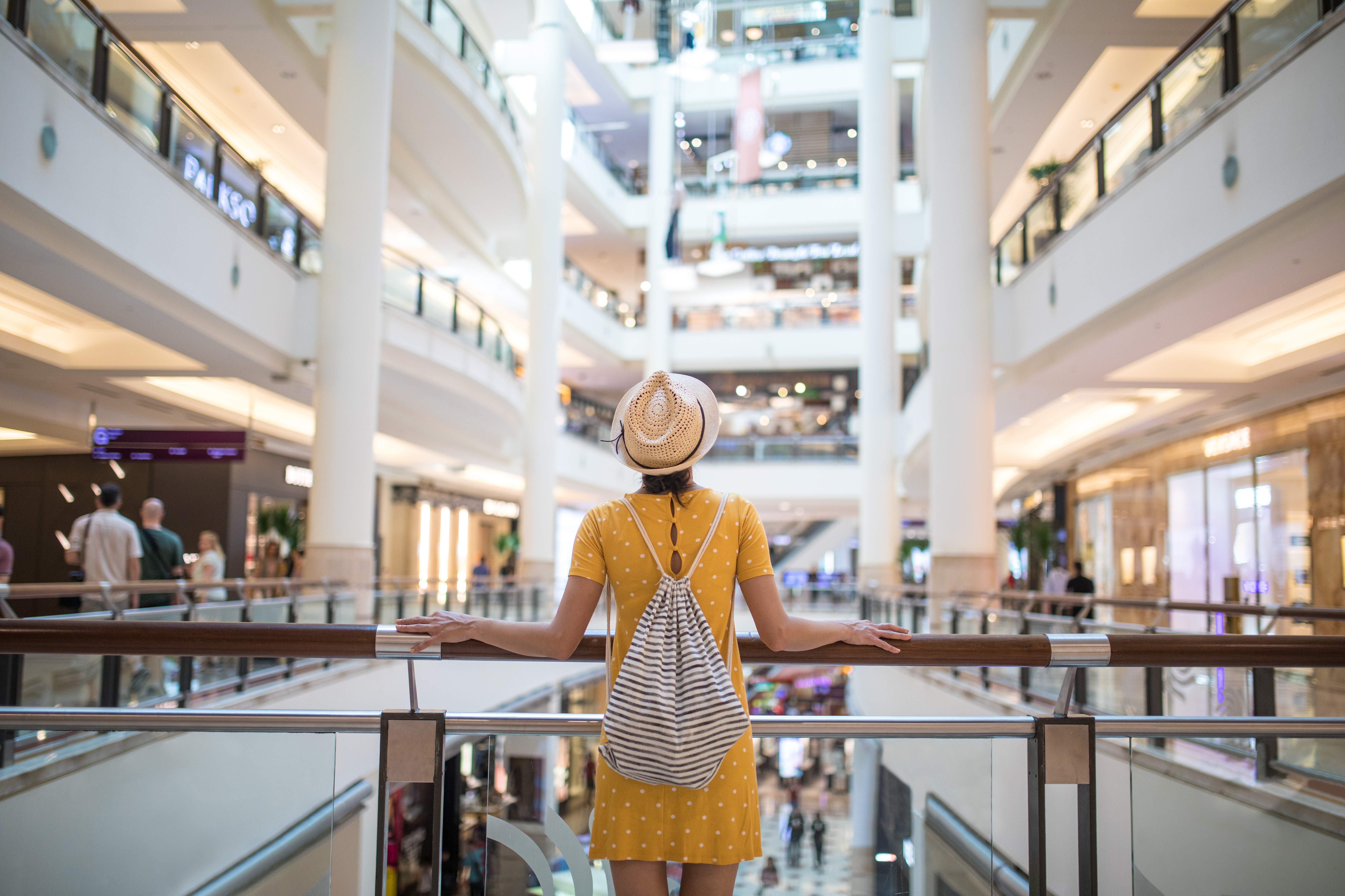 A woman in a yellow dress and hat stands by a railing in a spacious building.
