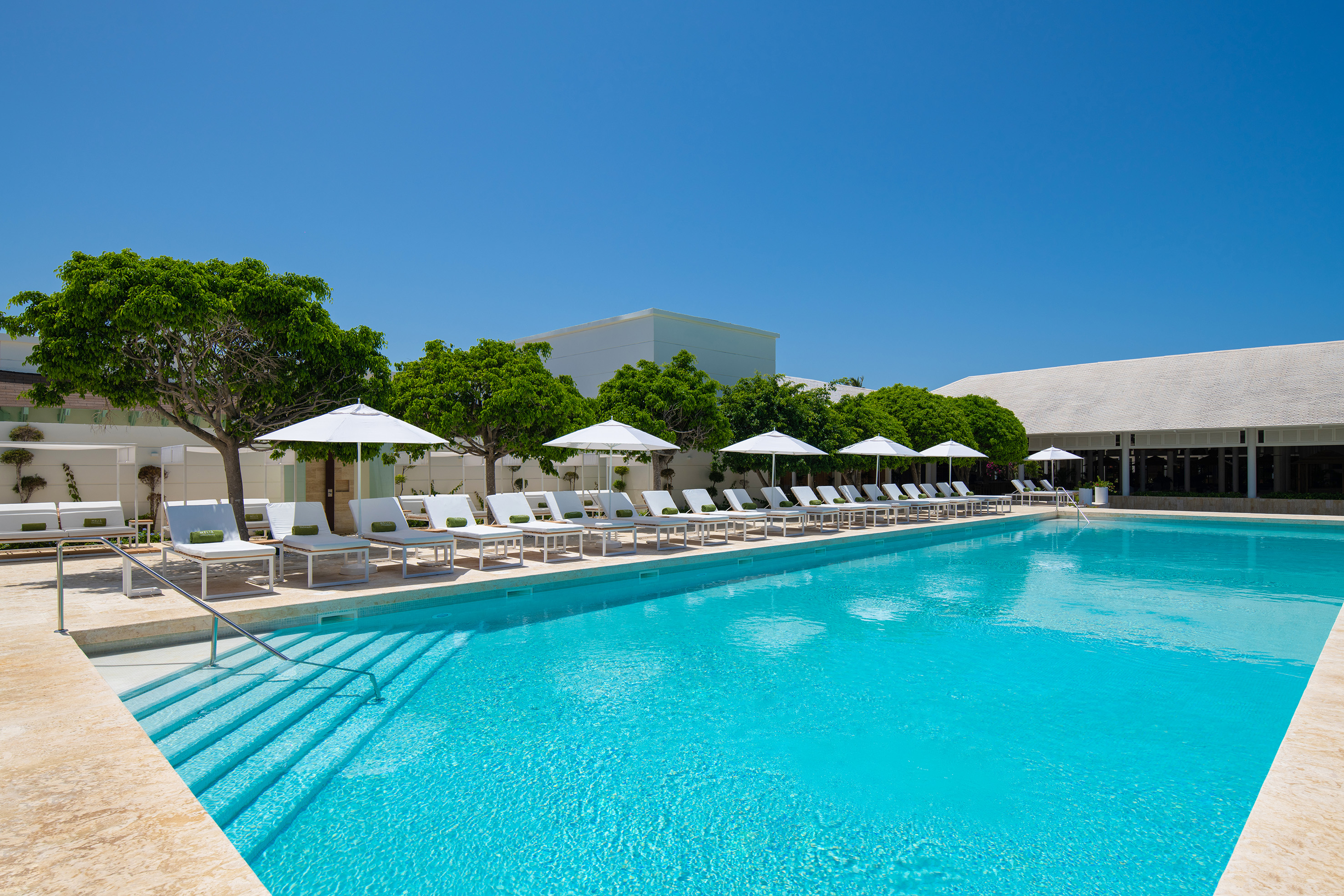 Resort swimming pool with white lounge chairs, umbrellas, and green trees.