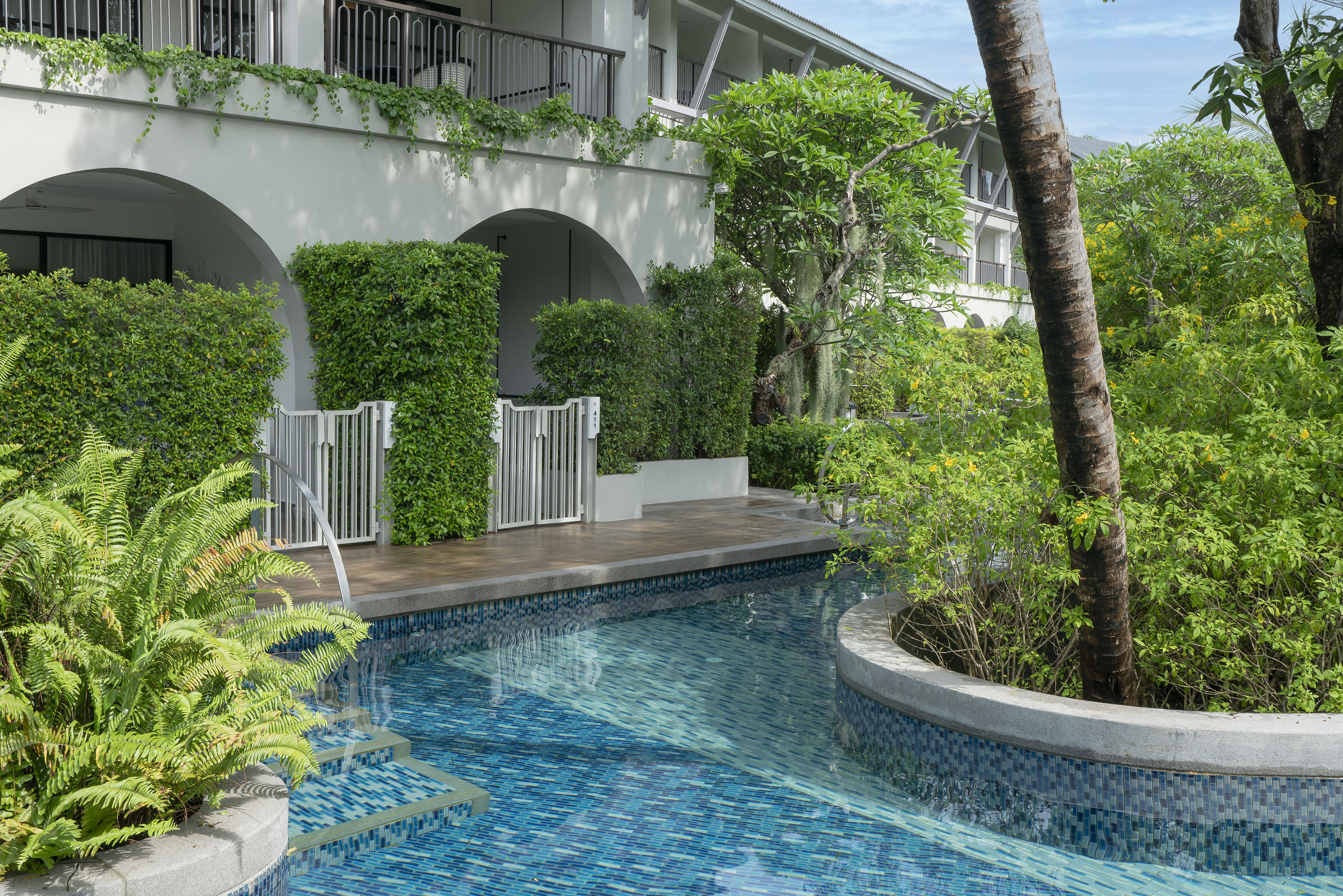 Tropical resort pool with blue mosaic tiles, green plants, and hotel architecture.
