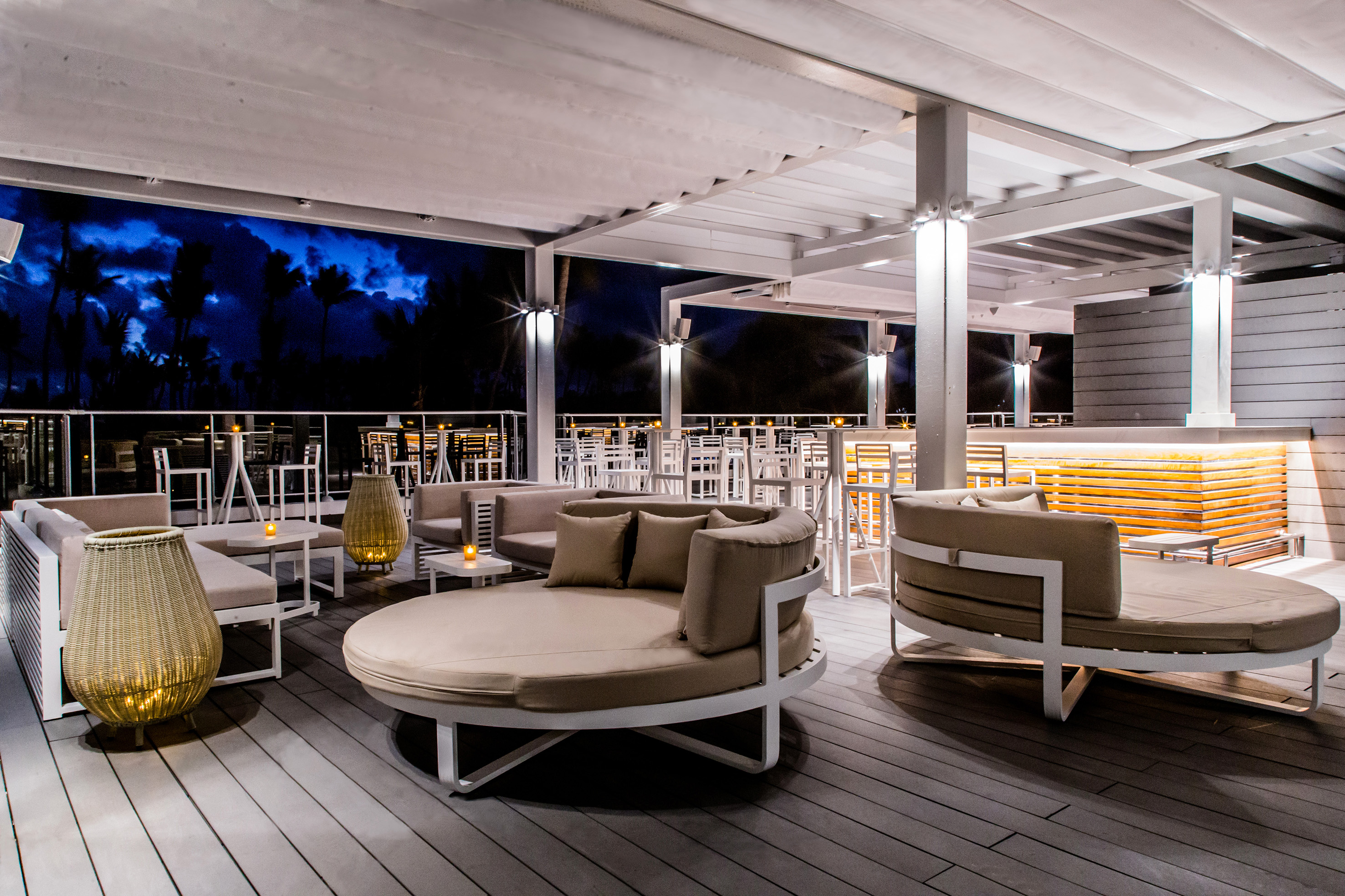 Rooftop bar and lounge at night with modern seating, illuminated bar, palm trees.