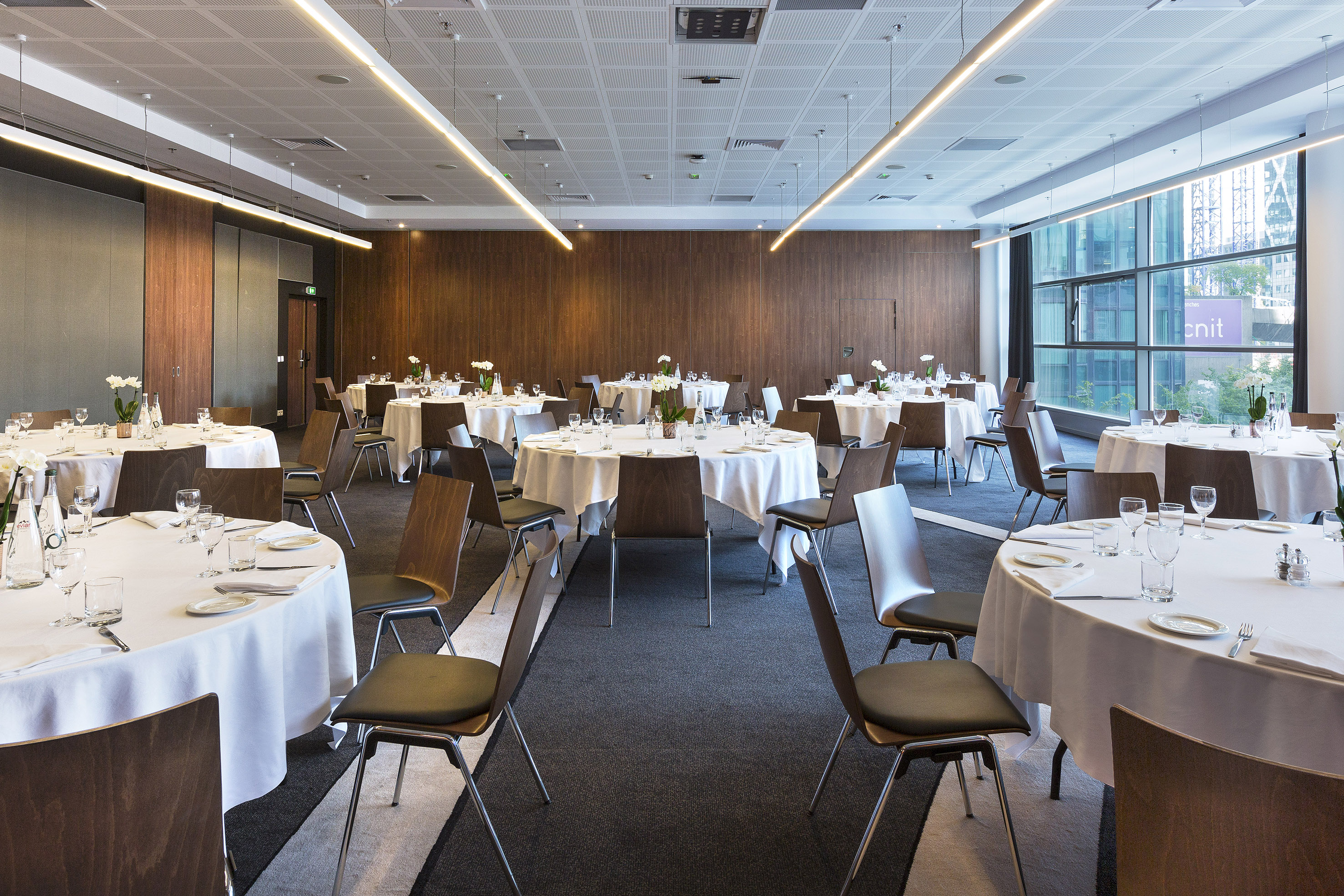 Modern banquet hall with round tables, white linens, wood chairs, and city views.