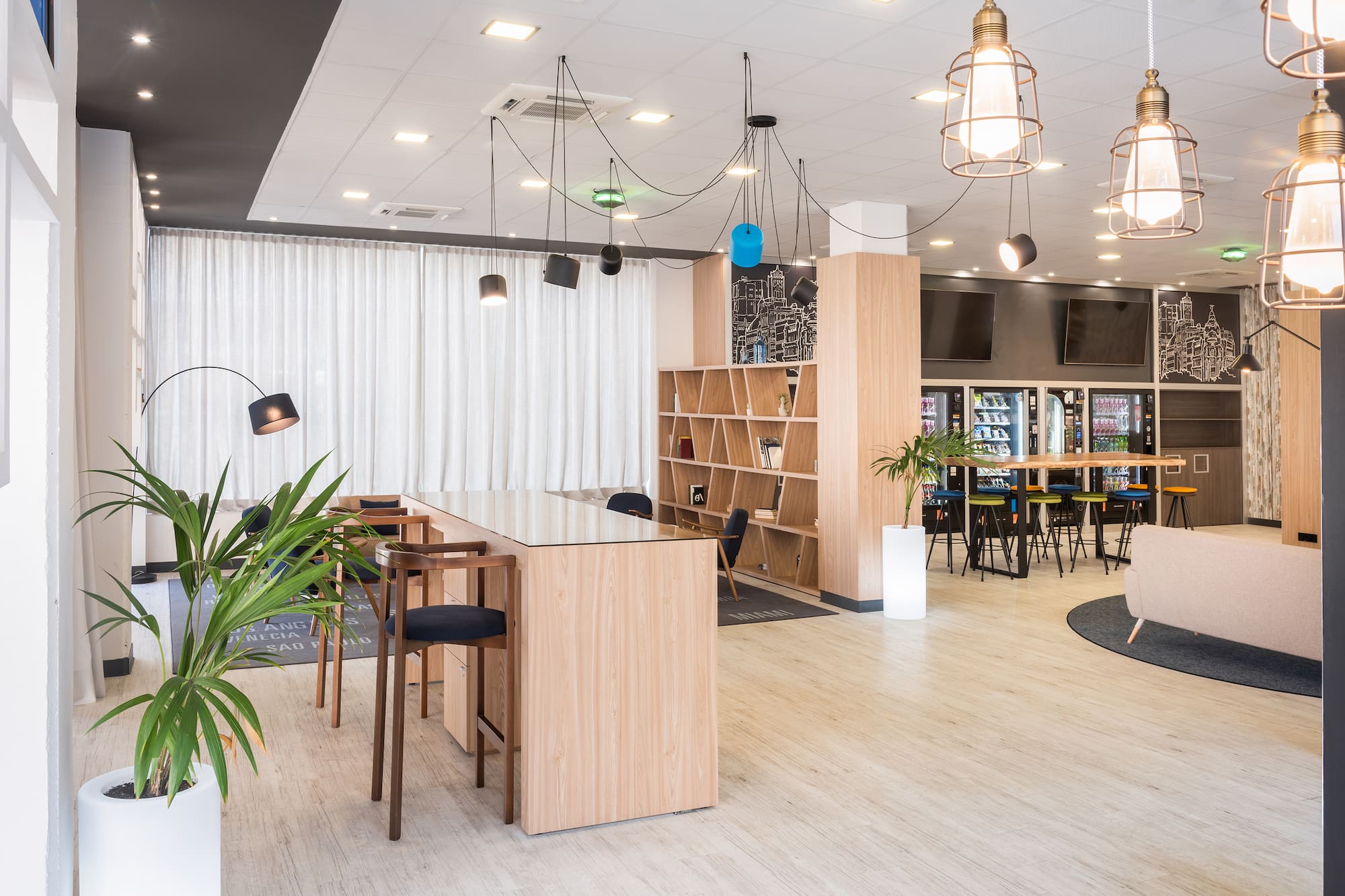Modern communal area with wooden tables, shelves, plants, and vending.