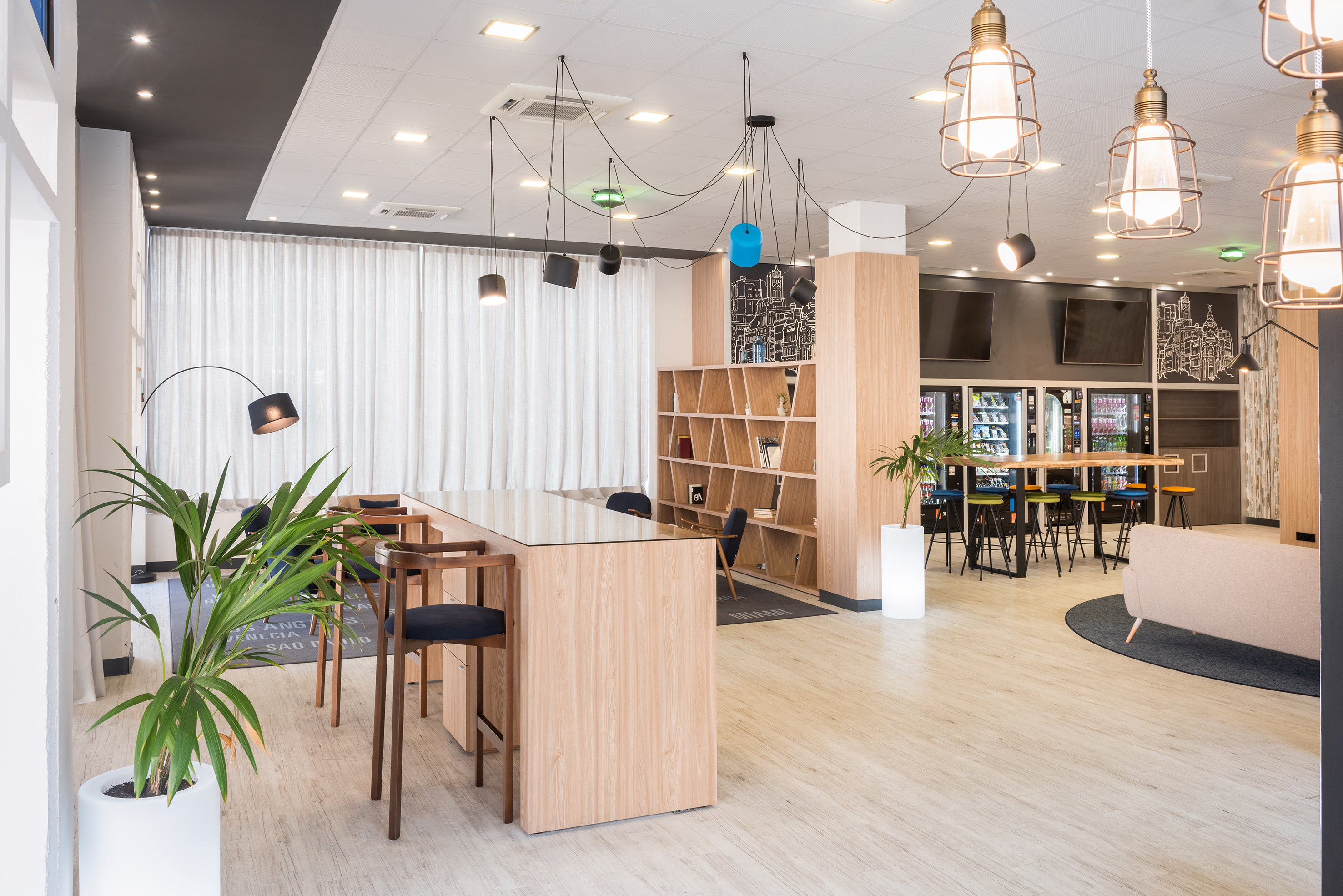 Modern communal area with wooden tables, shelves, plants, and vending.