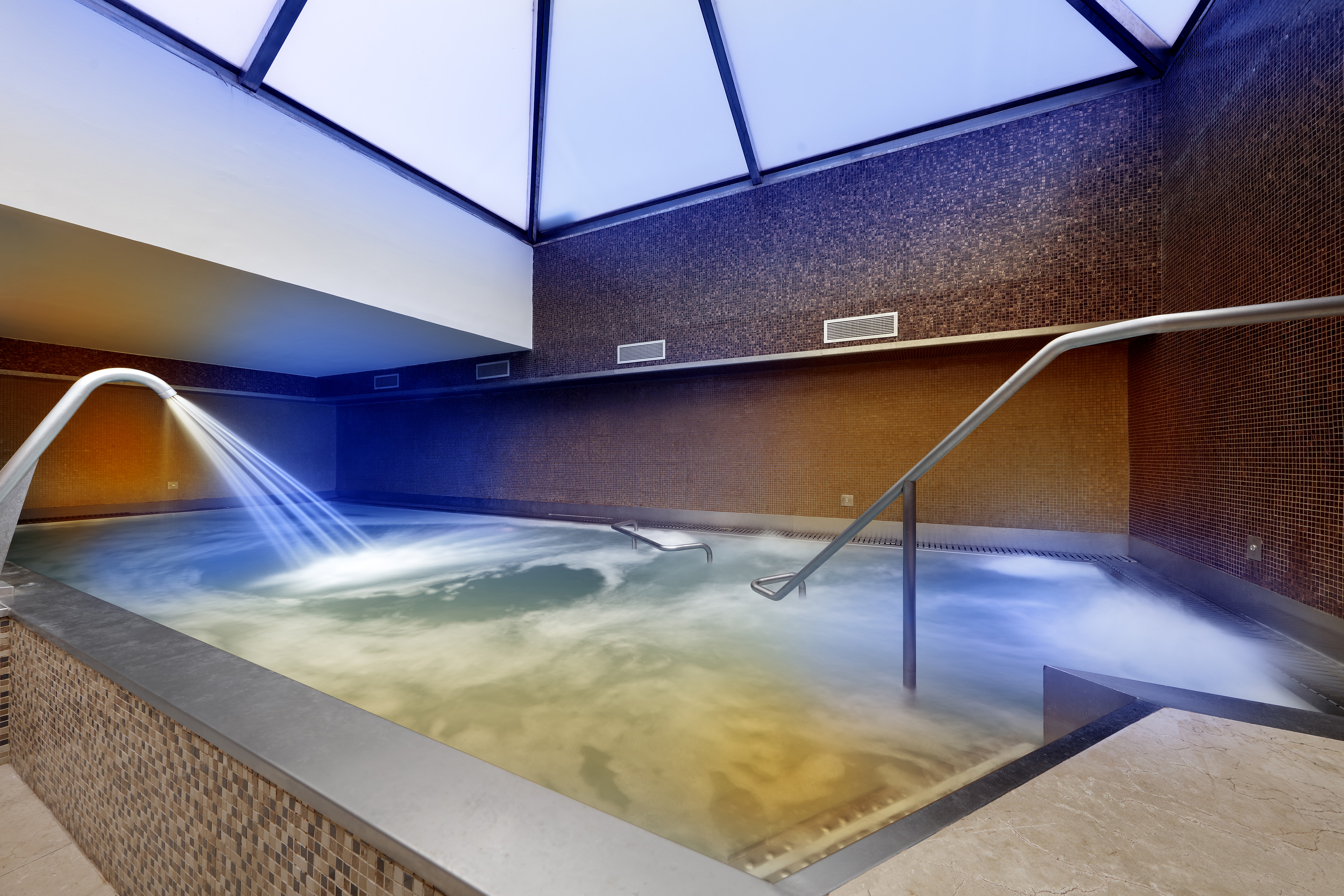Steaming indoor spa pool with water jet, mosaic tiles, and skylight.