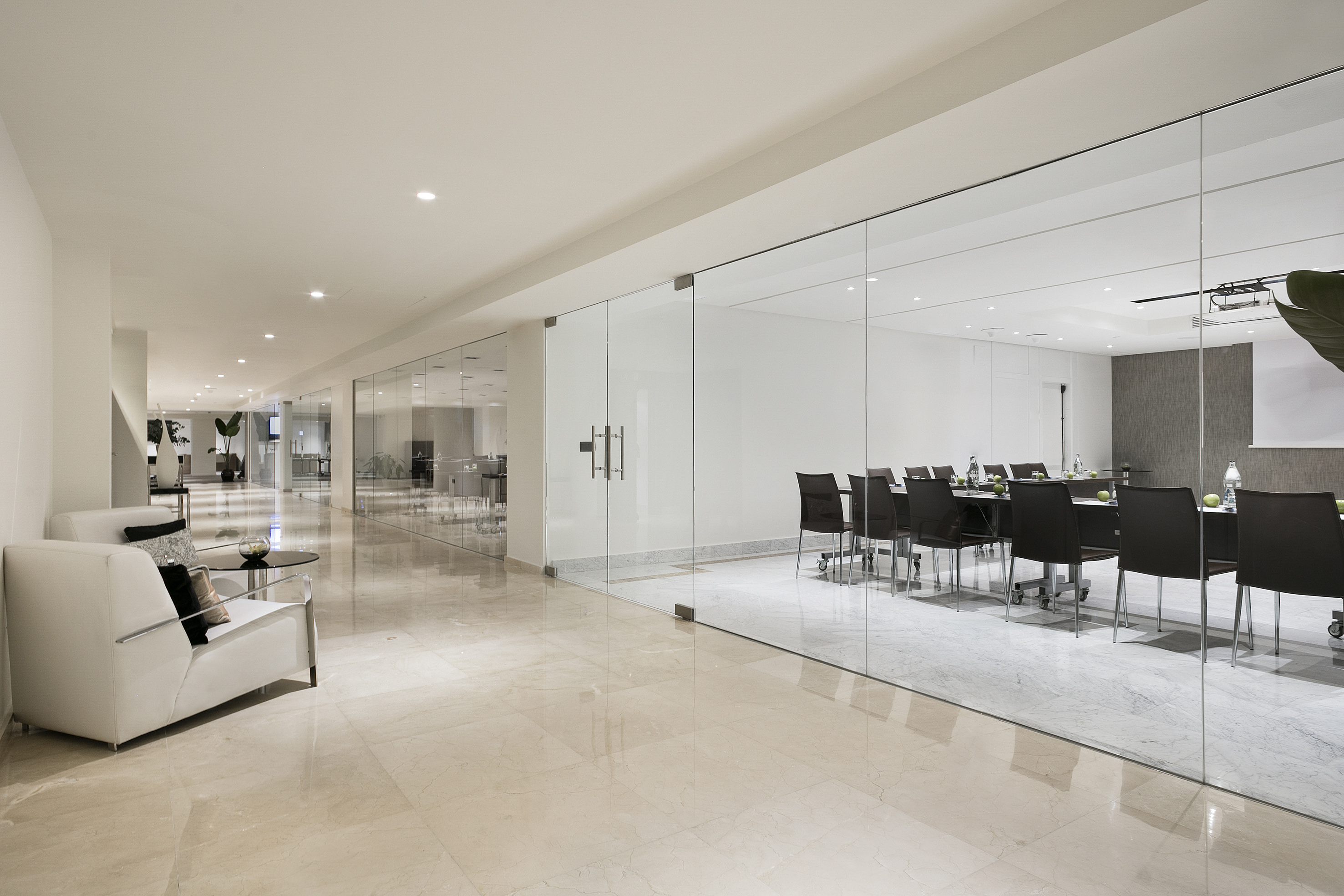 Modern hallway with glass meeting room, white lounge chairs, and marble floor.
