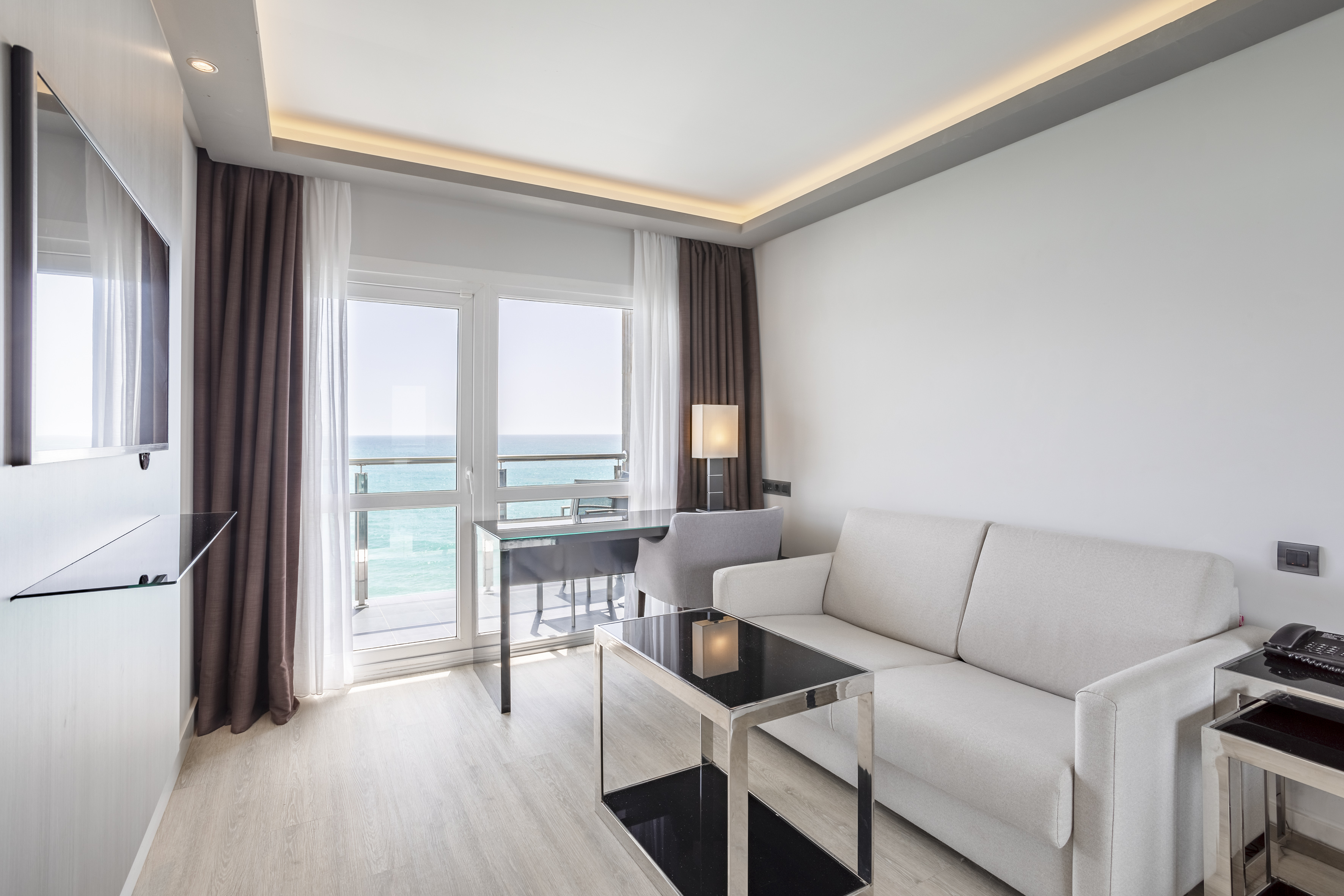 Modern hotel room with ocean view balcony, white sofa, and glass desk.