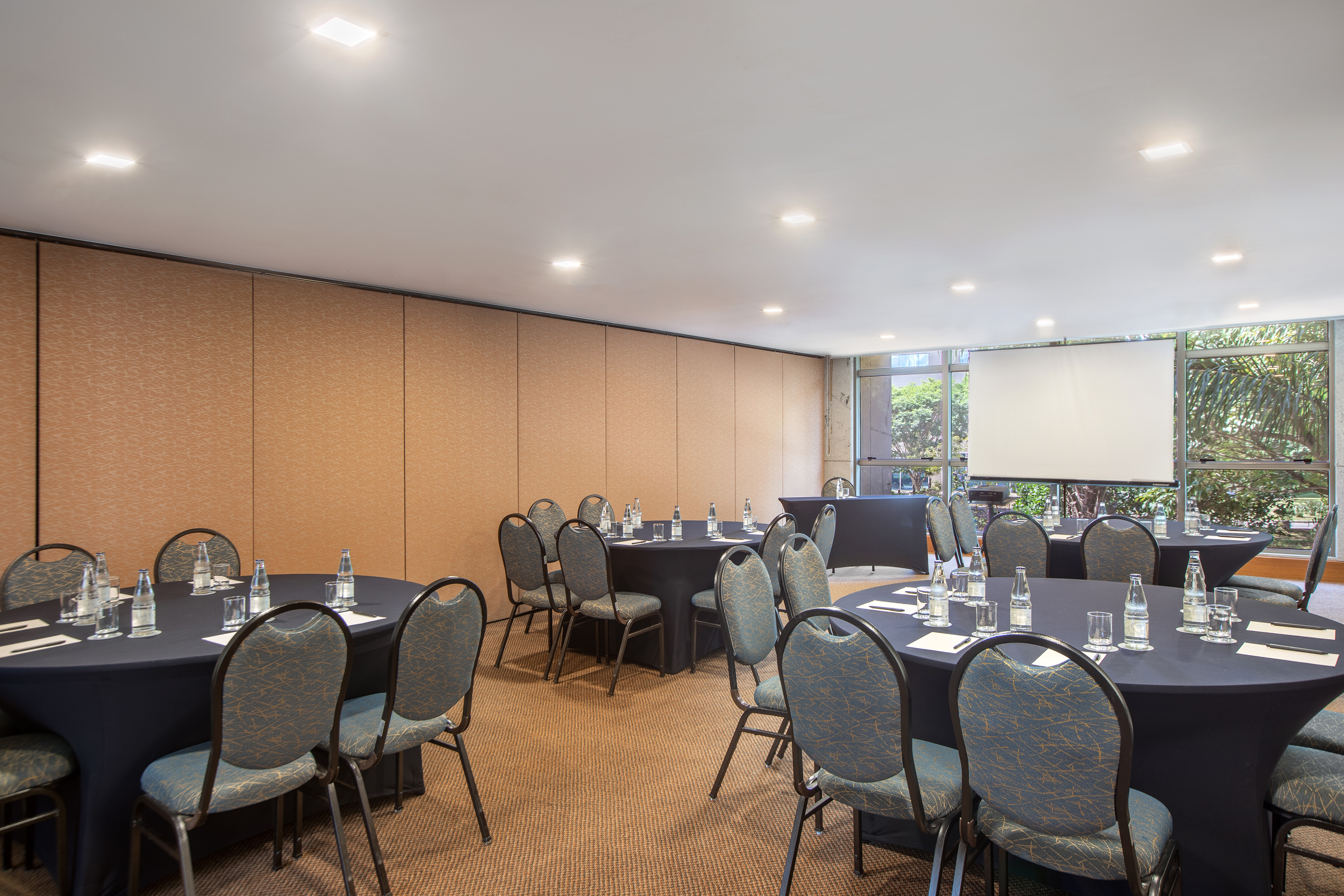 Modern meeting room with round tables, chairs, screen, and natural light. - Encontro I - -Hotel Madrid Airport Suites Affiliated by Meliá:, Brasilia