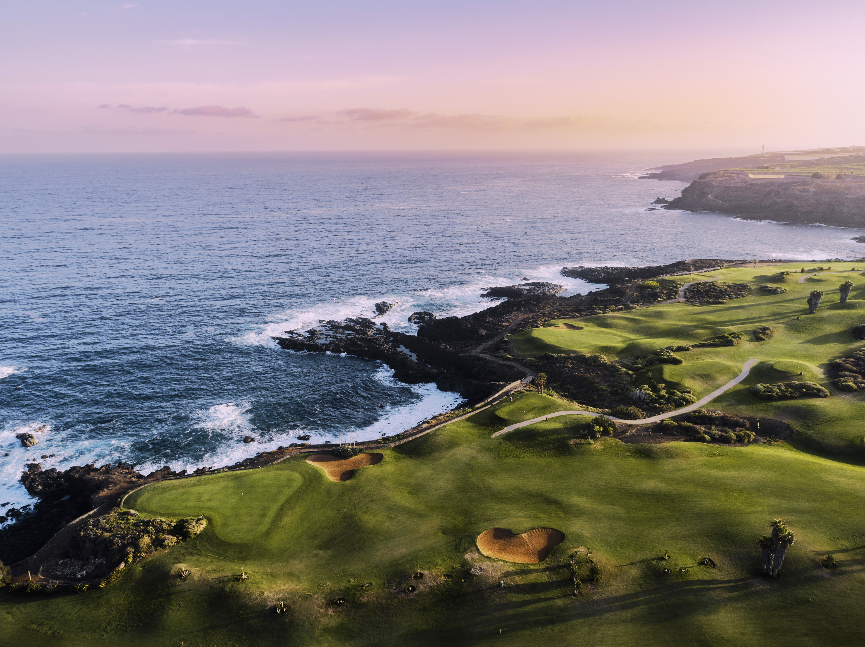 Aerial view of coastal golf course with ocean, green fairways, sand bunkers, sunset sky.