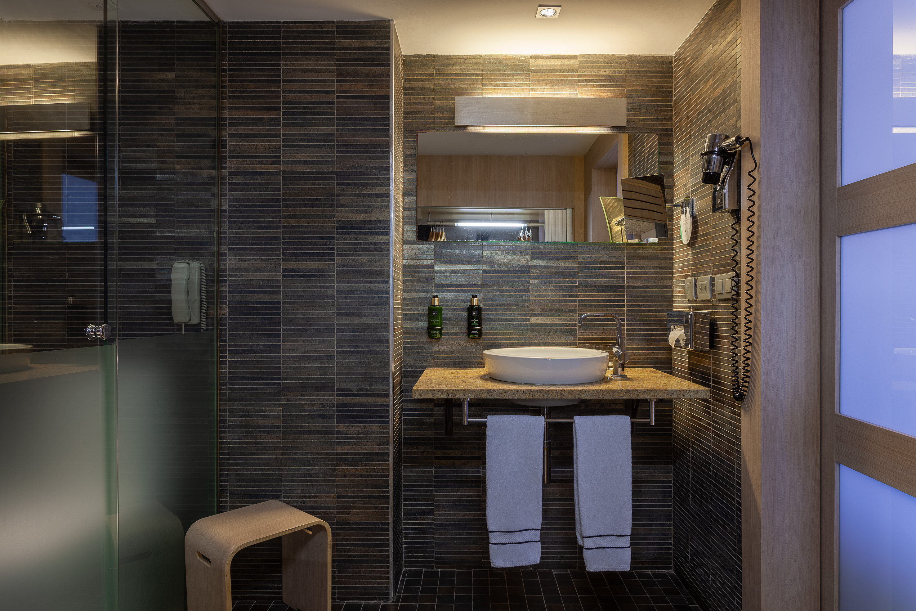 Hotel bathroom with dark tile, vessel sink, toiletries, and glass shower.