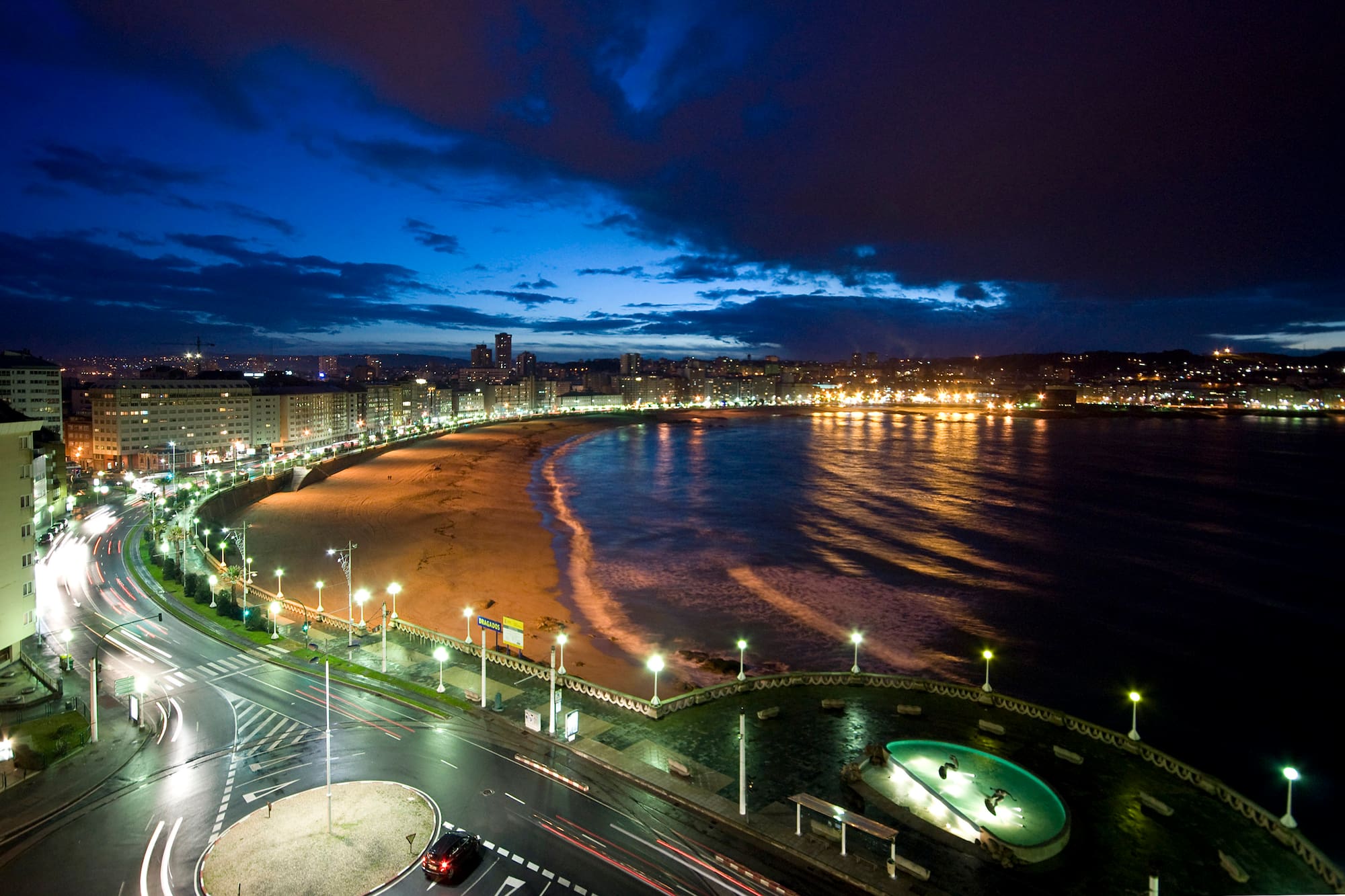 Panorámica nocturna de ciudad y bahía con playa, luces y estelas de coches.