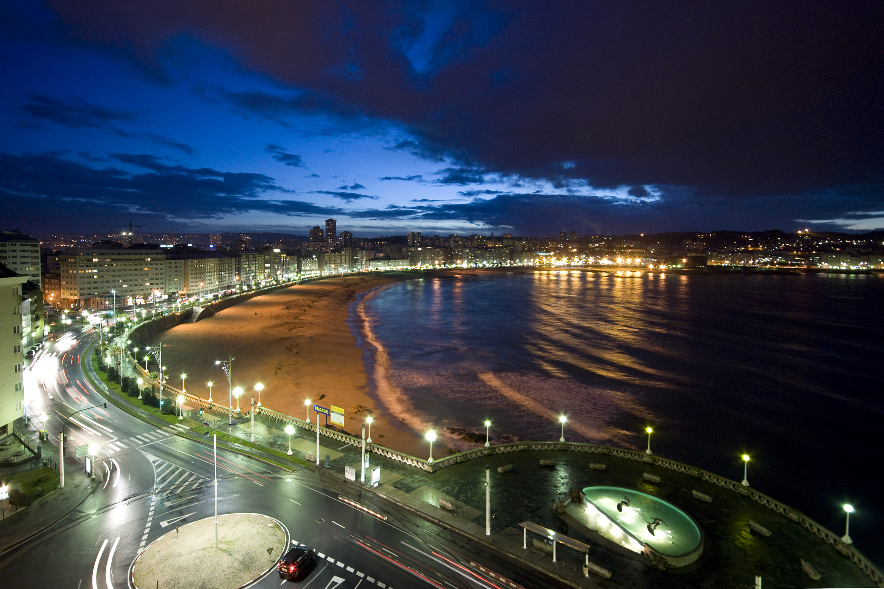 Panorámica nocturna de ciudad y bahía con playa, luces y estelas de coches.