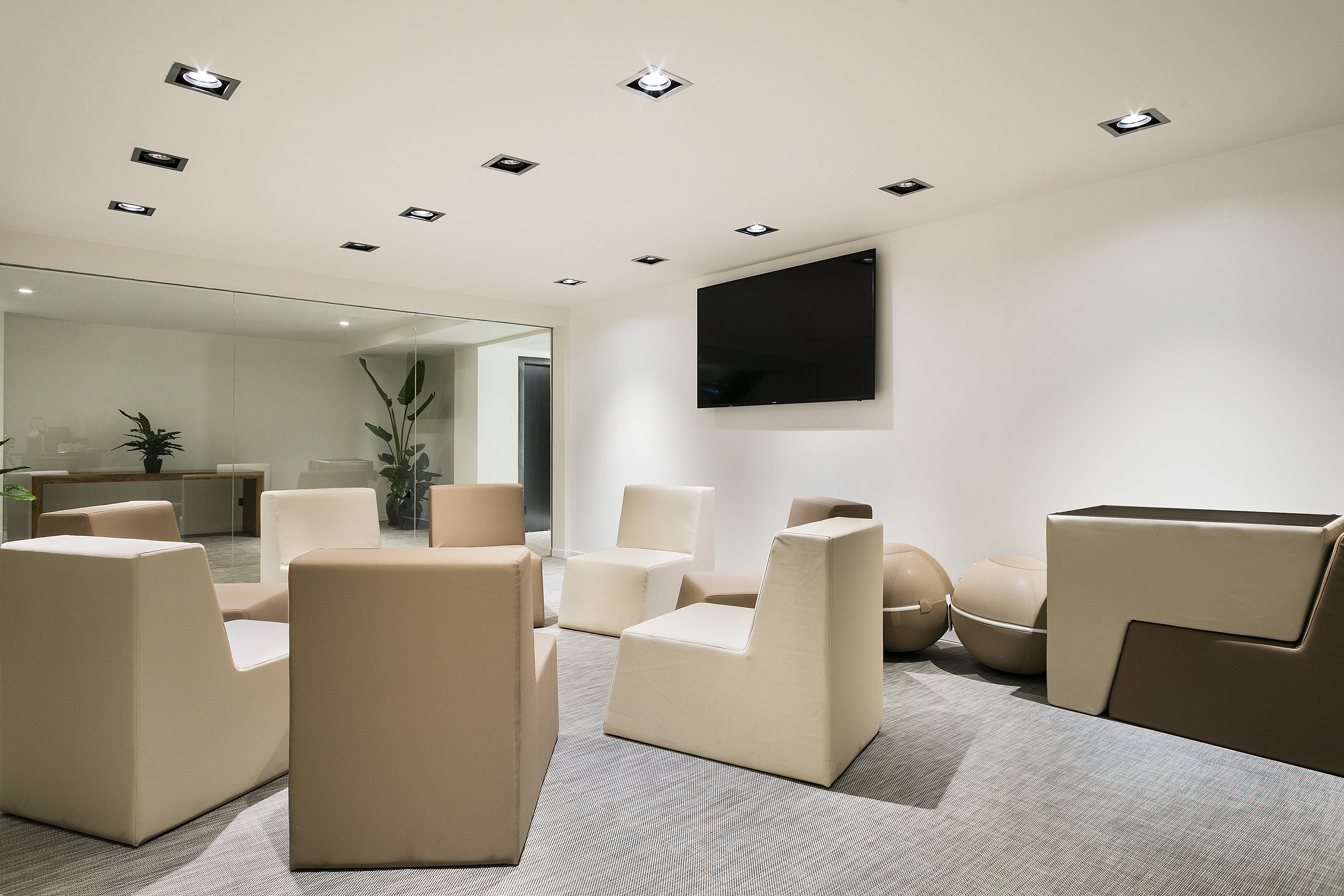 Modern office lounge with neutral-toned seating, recessed lighting, and TV.