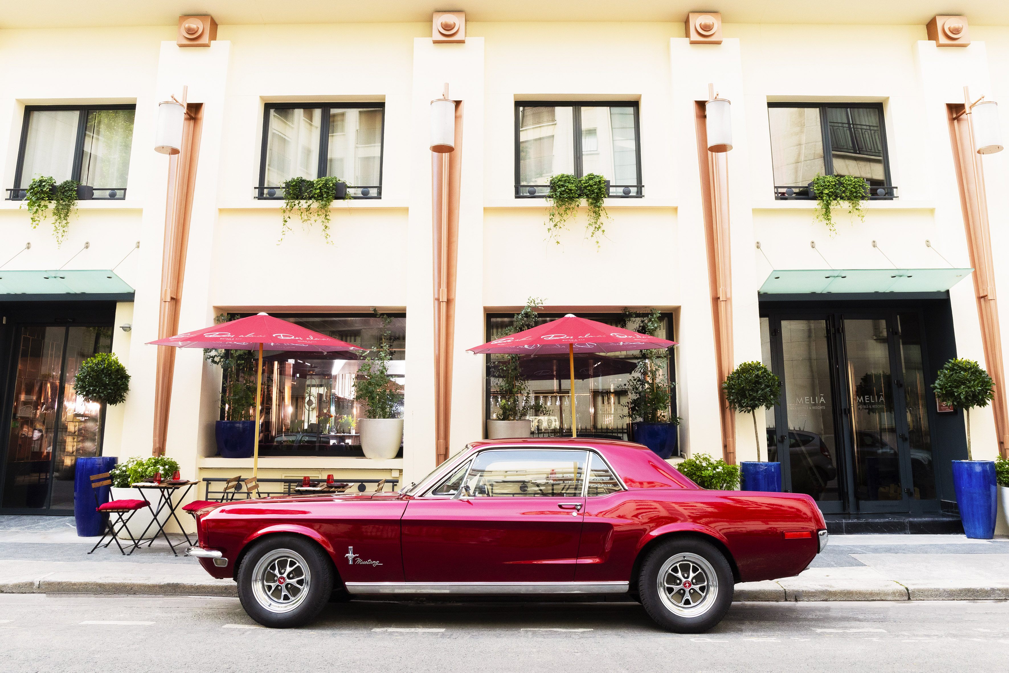 Red vintage Mustang parked outside Melia hotel cafe with umbrellas.