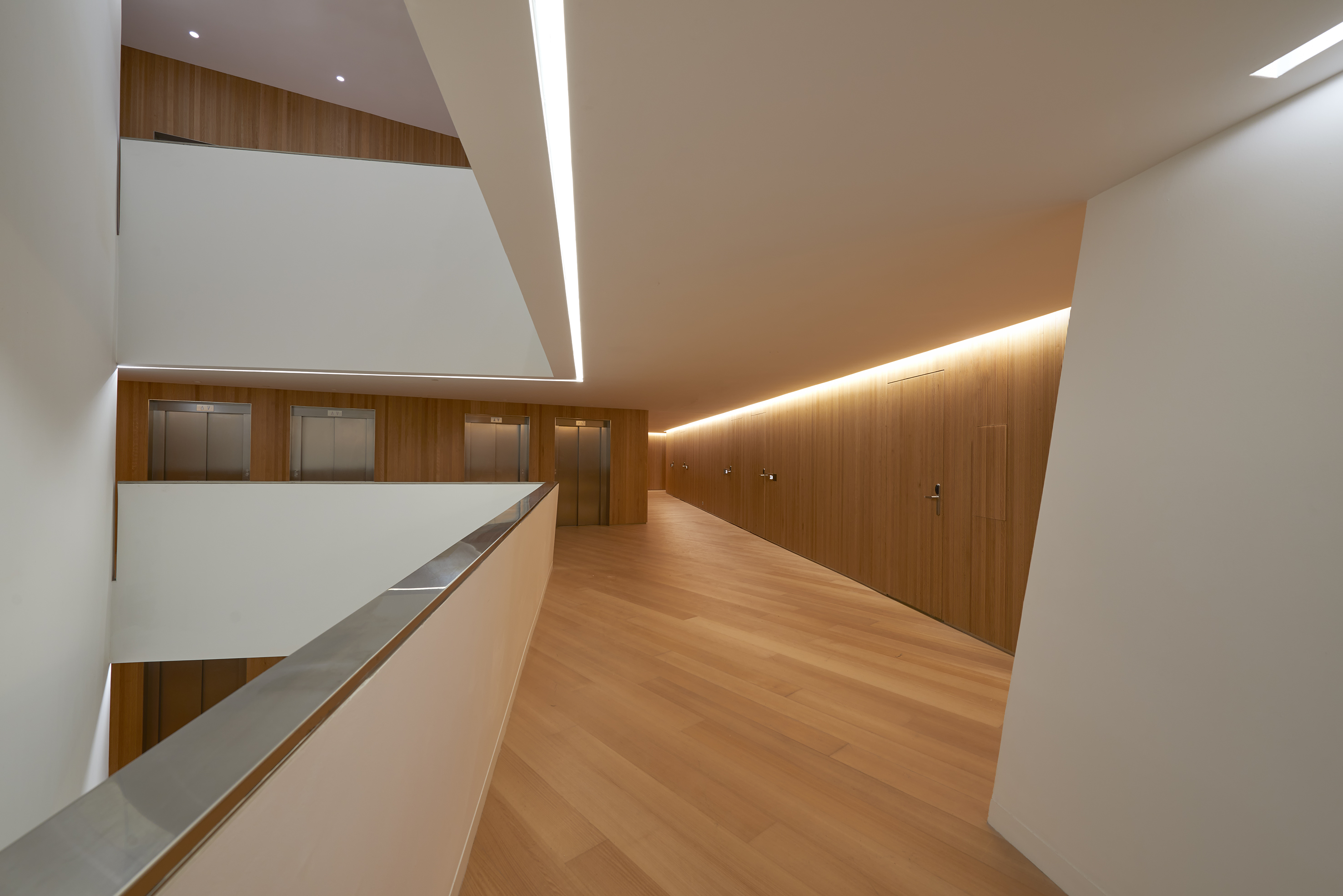 Modern wood-paneled hallway with linear lighting and multiple stainless steel elevators.