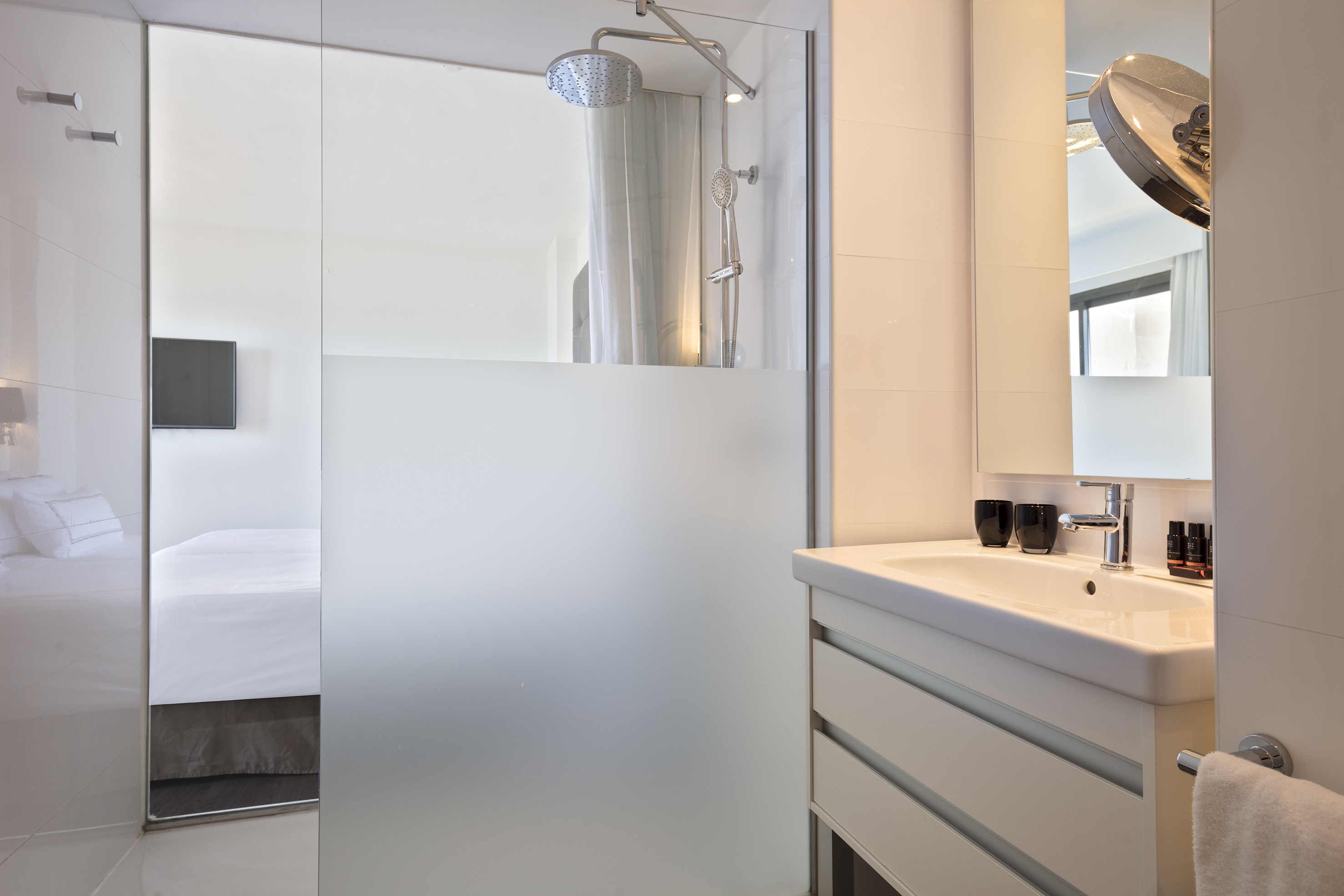 Modern bathroom with glass shower, rain head, white vanity, and hotel bedroom.