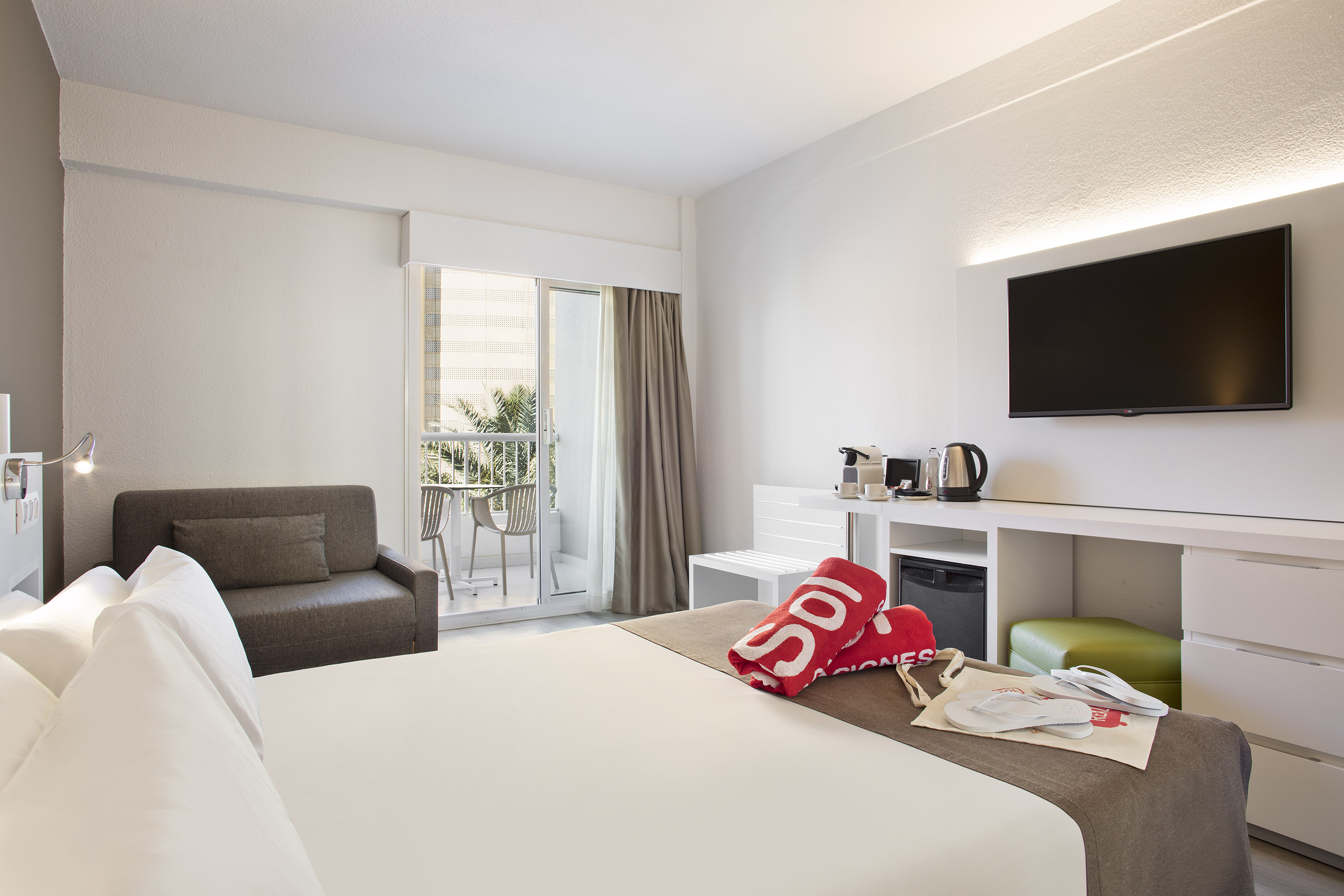 Bright hotel room with bed, sofa, balcony access, TV, and coffee station.