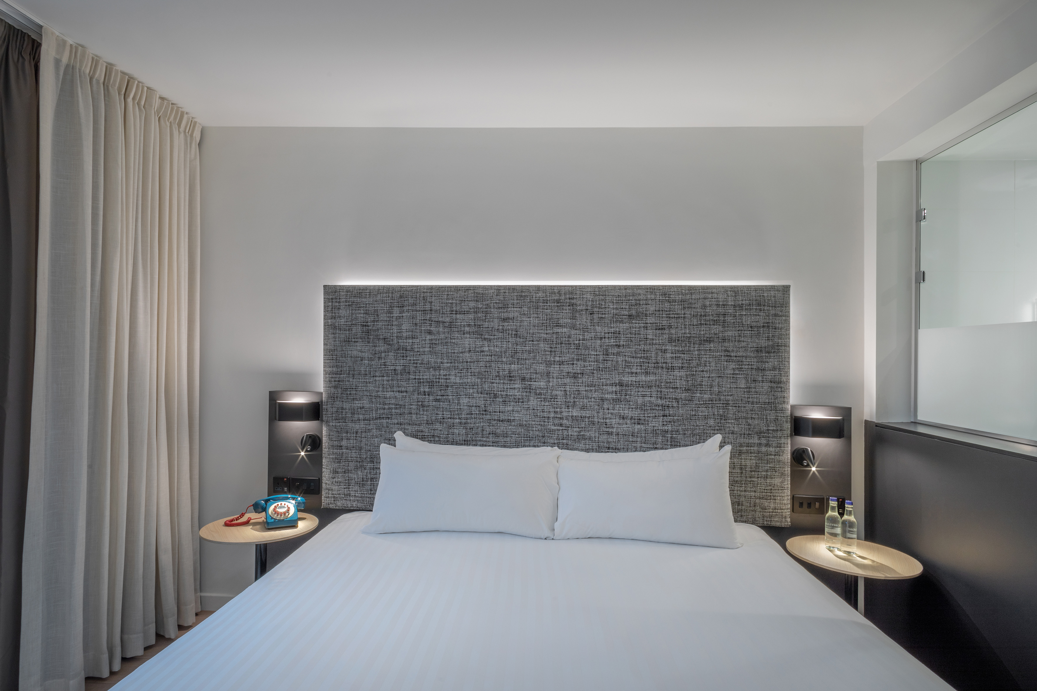 Hotel guest room with white bed, grey headboard, bedside tables, and ambient lighting.