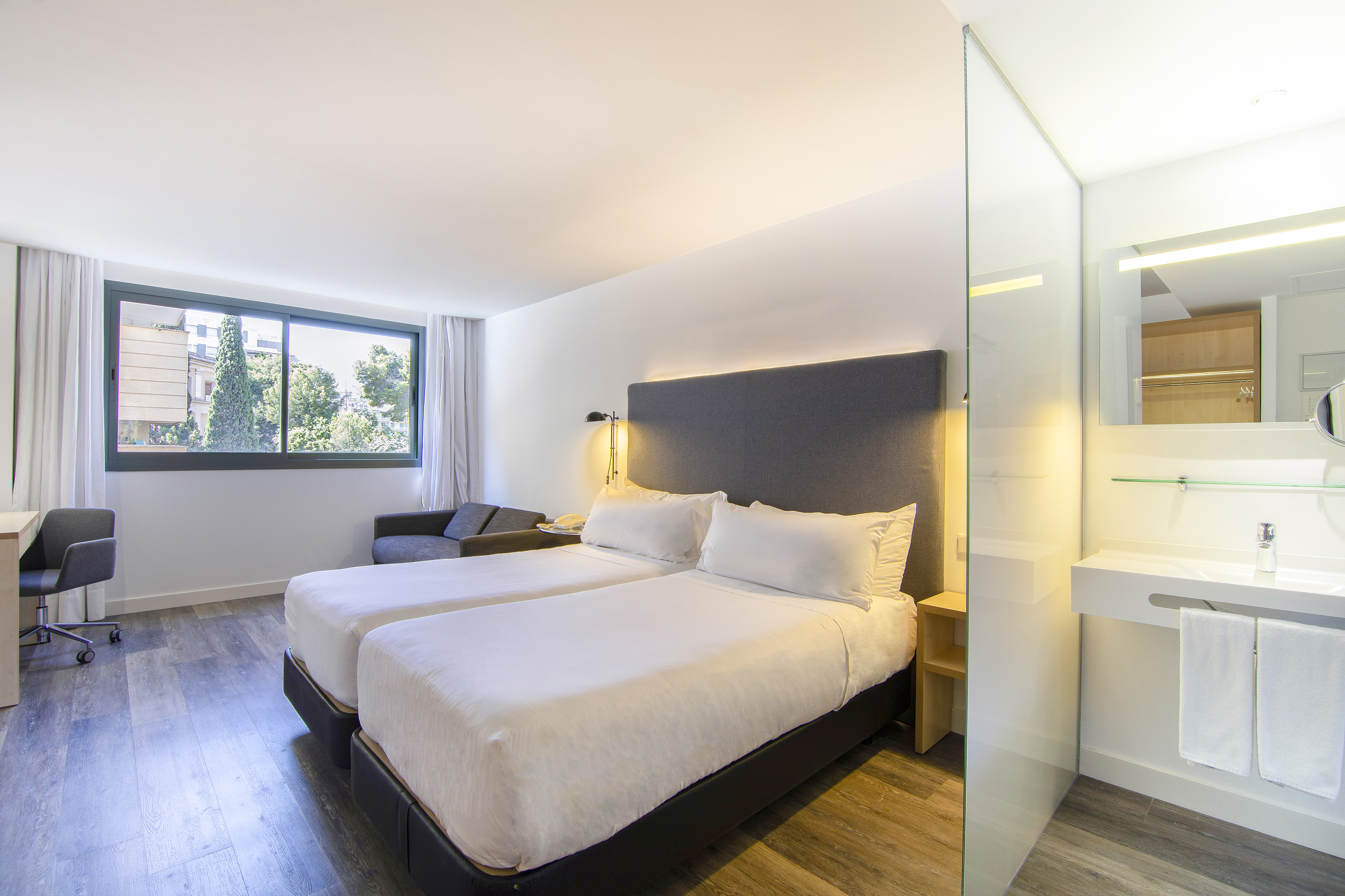 Modern hotel room with two twin beds, desk, and glass-partitioned bathroom.