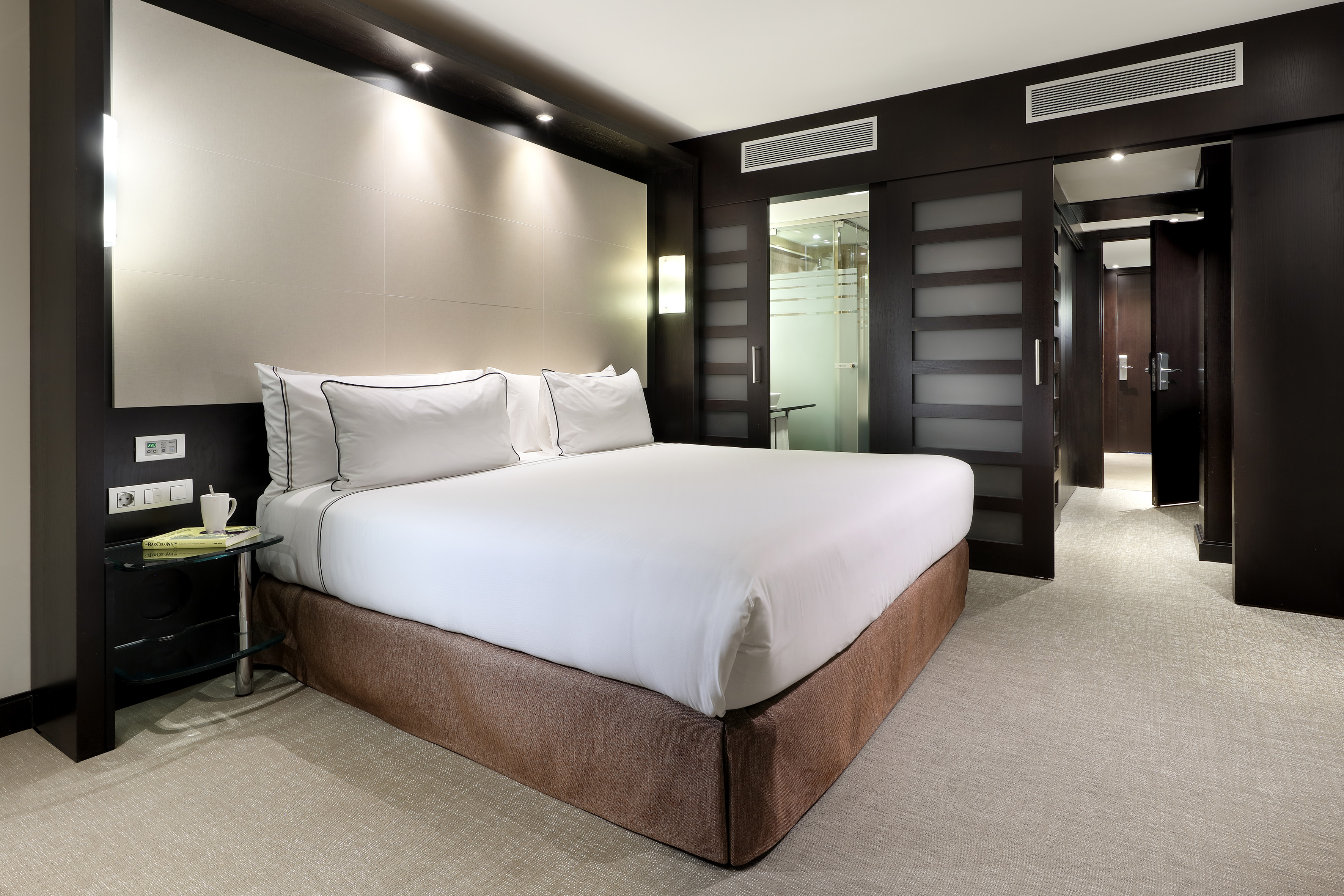 Modern hotel bedroom with white bed, dark wood decor, and glass sliding doors.