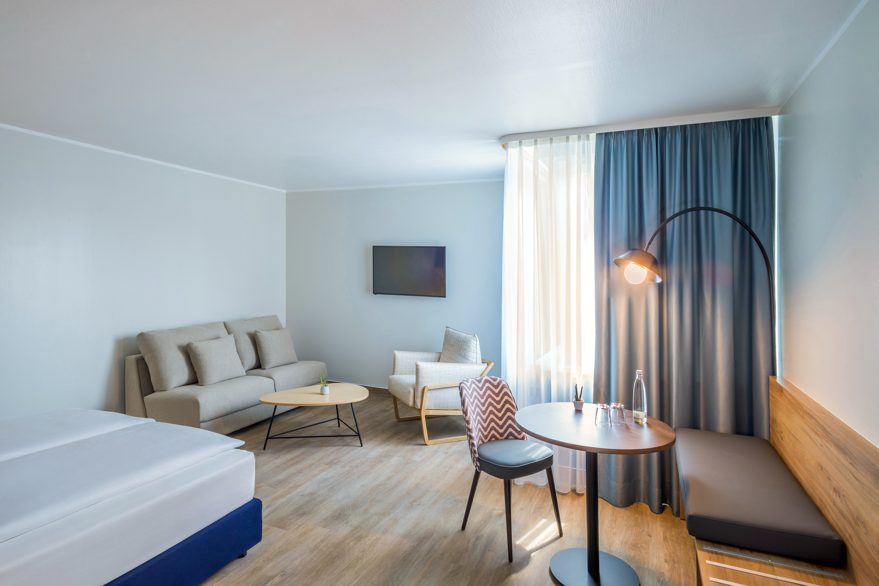 Modern hotel suite with bed, sofa, TV, dining area, and blue curtains.
