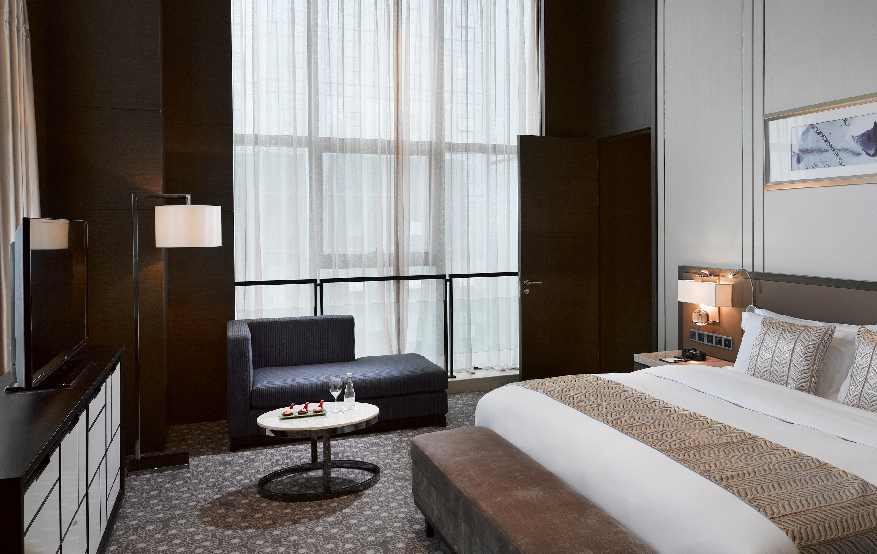 Luxury hotel room with king bed, modern sofa, and window with sheer curtains.