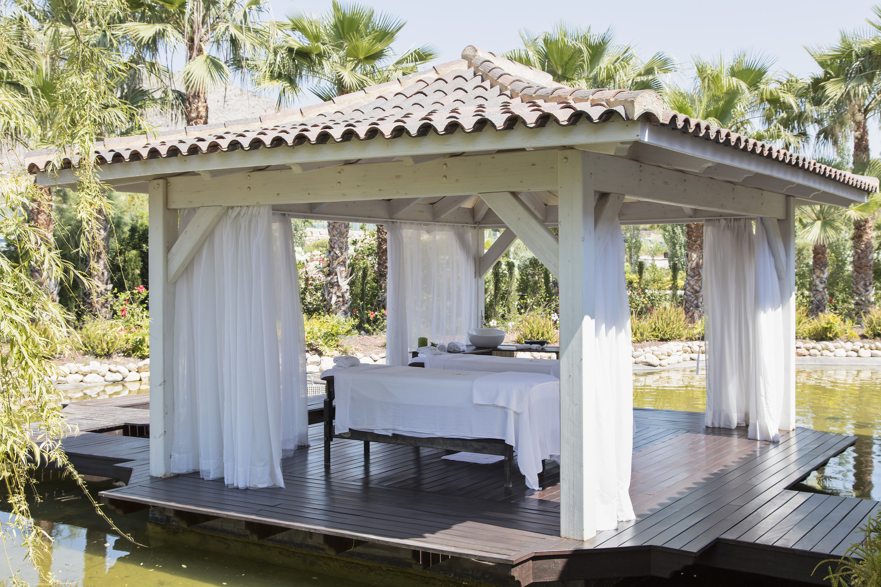 Tropical overwater spa gazebo with massage bed, white curtains, palm trees.