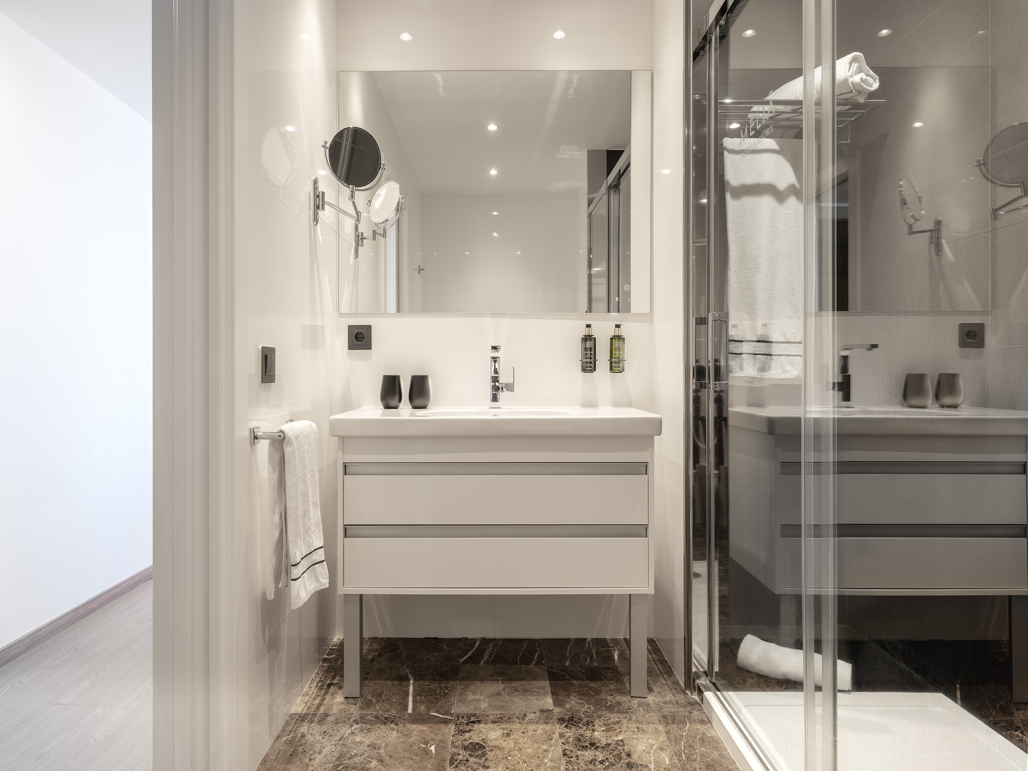 Modern bathroom with white vanity, large mirror, glass shower, and marble floor.