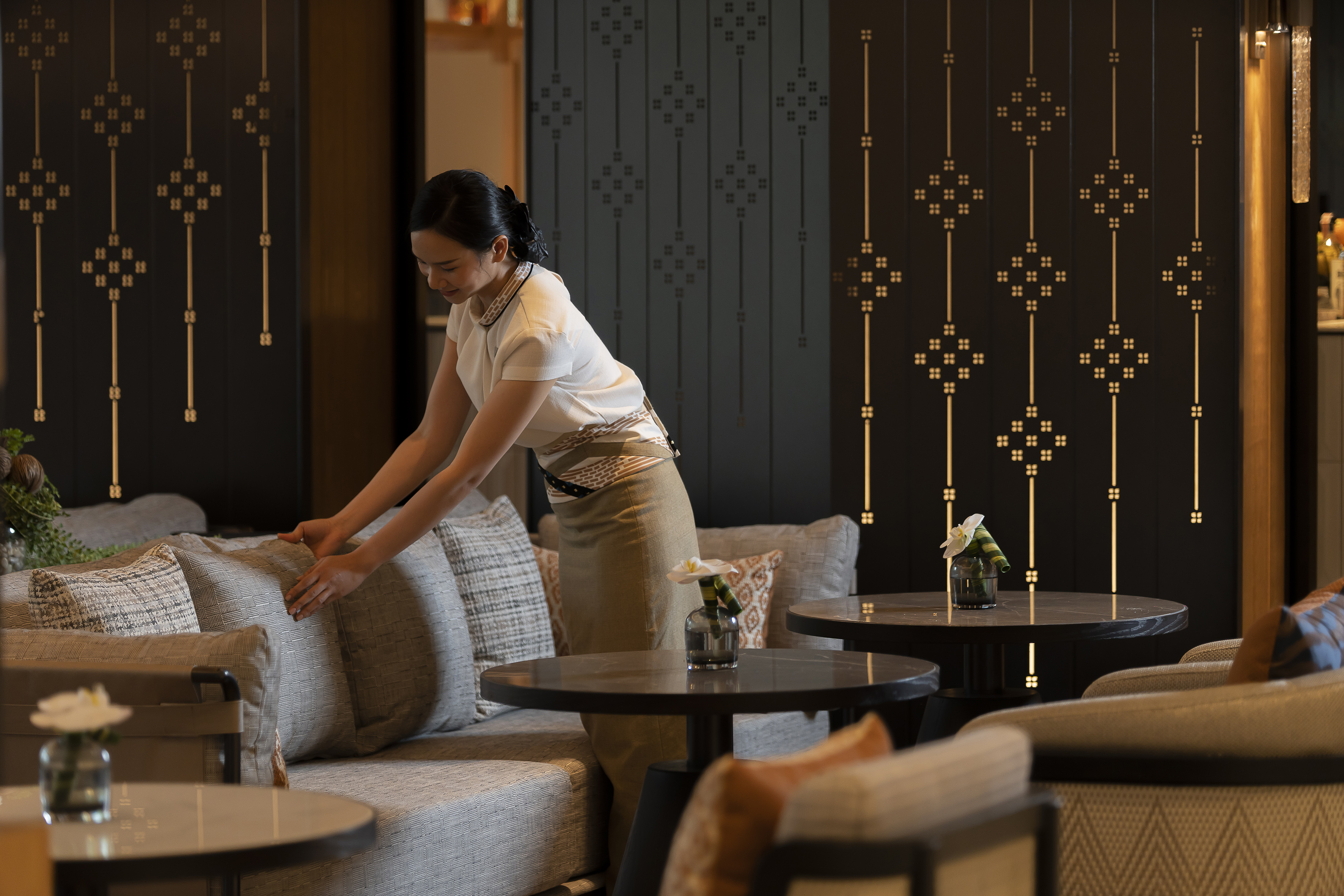 Attentive hotel staff arranges cushions on a sofa in a sophisticated lobby.