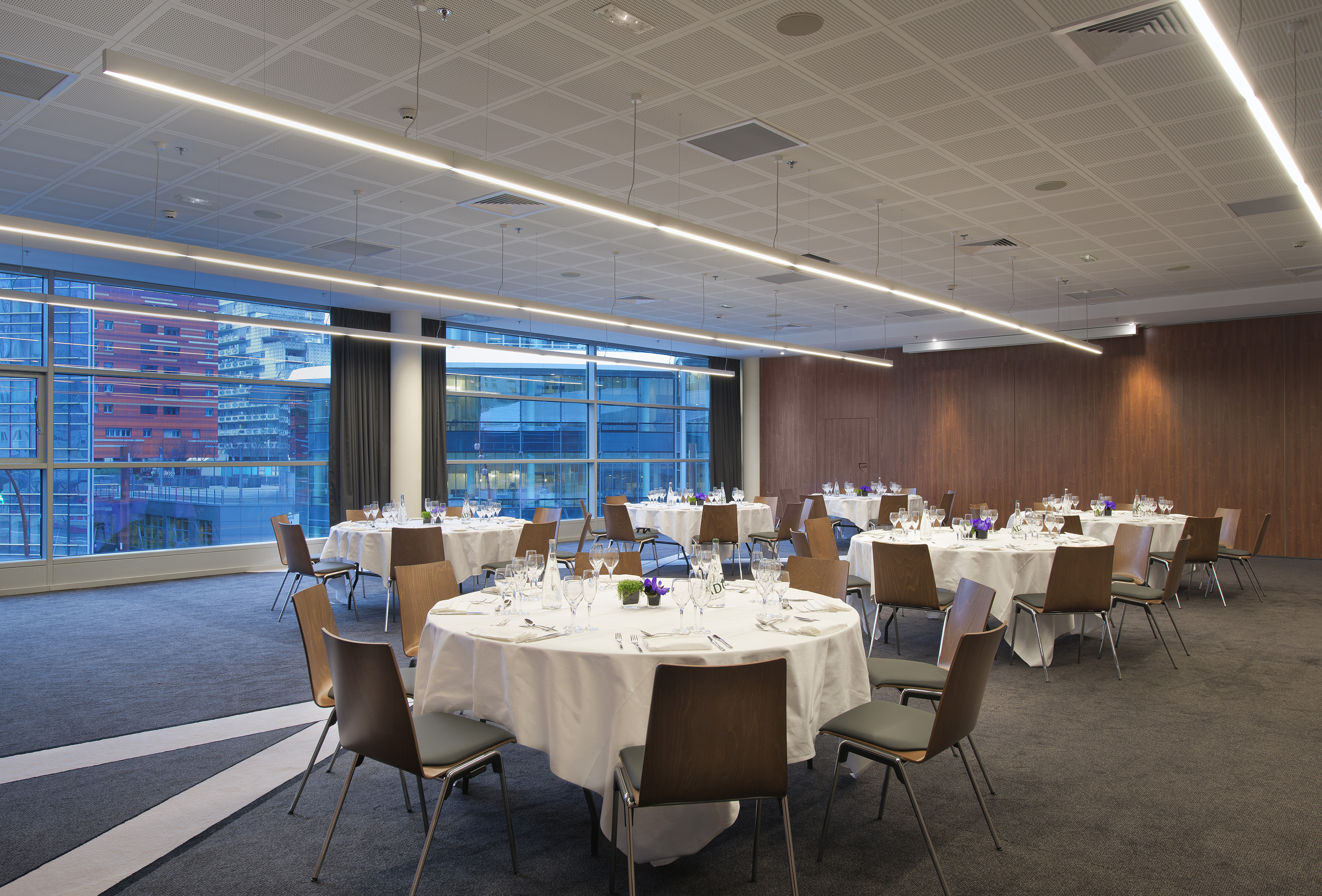 Modern banquet hall with round tables, place settings, city view windows.
