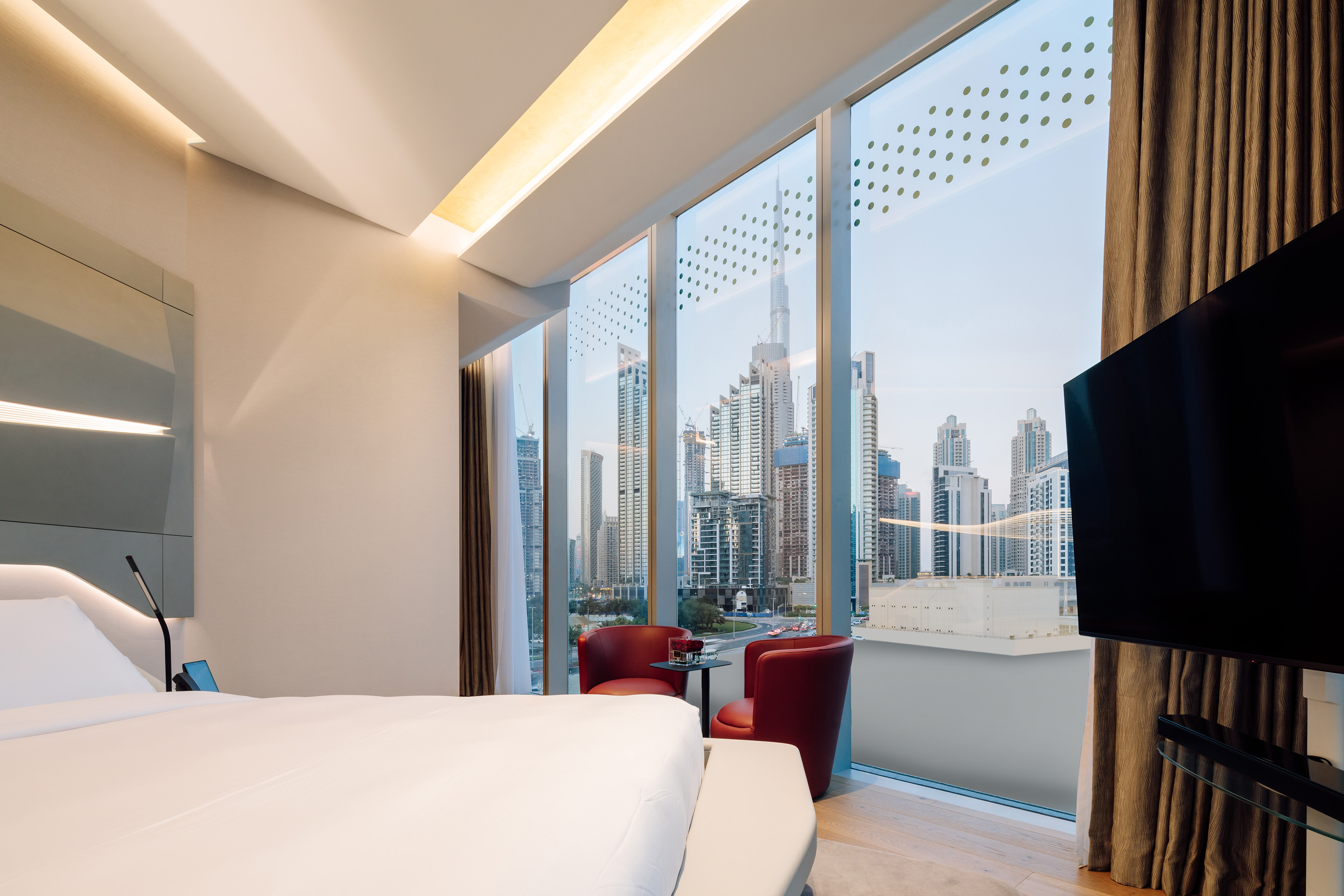 Modern hotel room with white bed, red armchairs, and Dubai city skyline view.