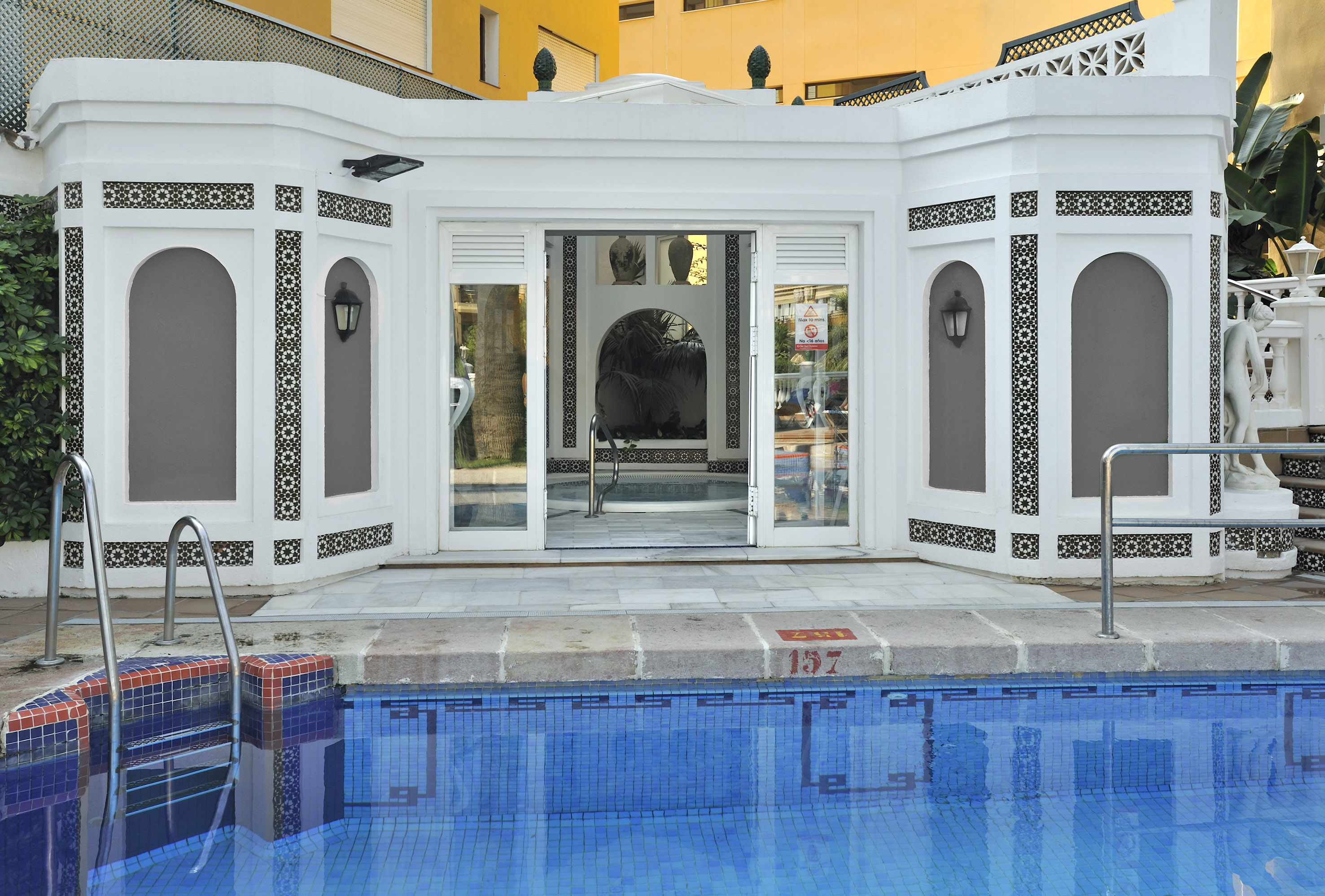 Blue pool, white building with intricate Moroccan tiles, leading to a spa area.
