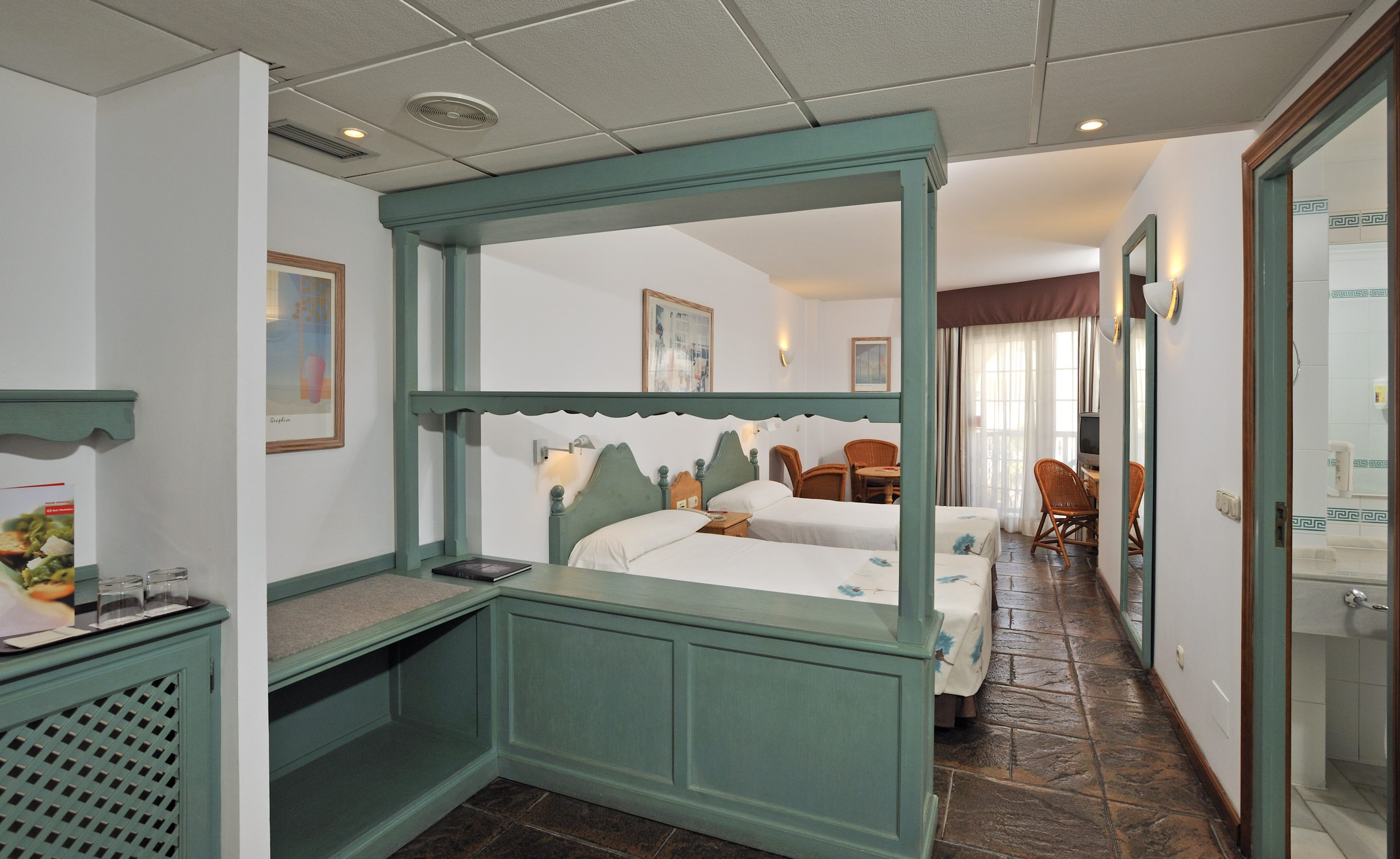 Hotel room with two twin beds, teal partition, and bright sitting area.