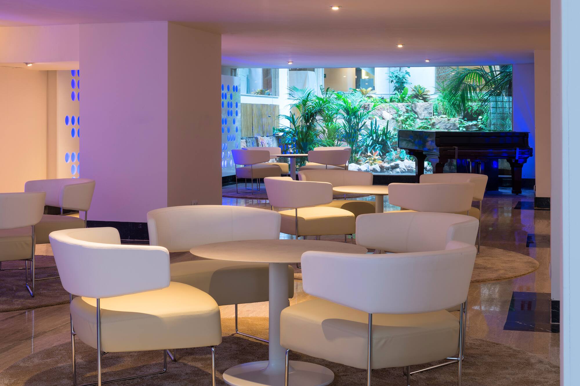 Modern hotel lounge with white chairs, round tables, lush indoor garden, and grand piano.