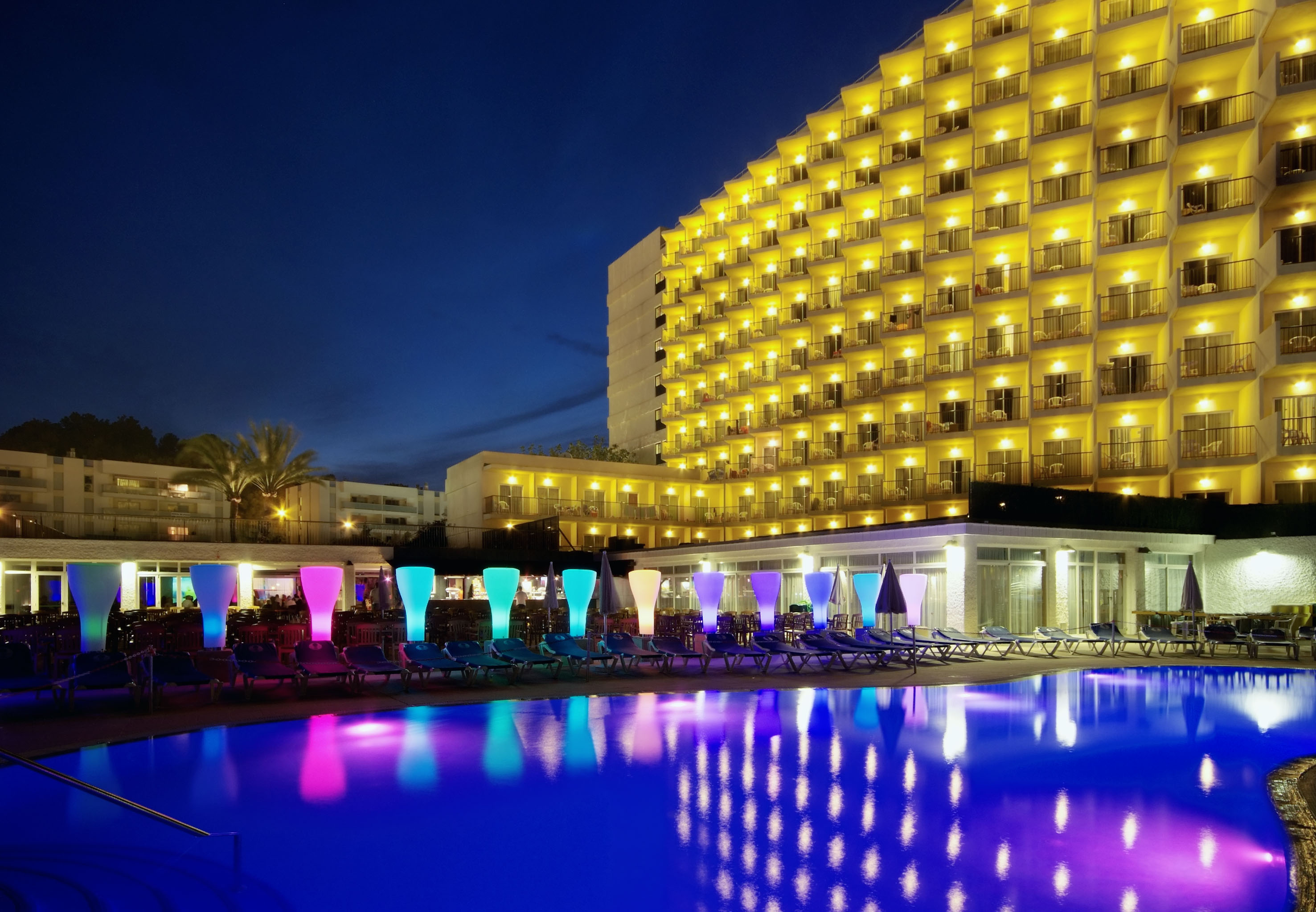 Illuminated resort hotel with colorful pool and lounge chairs at night.
