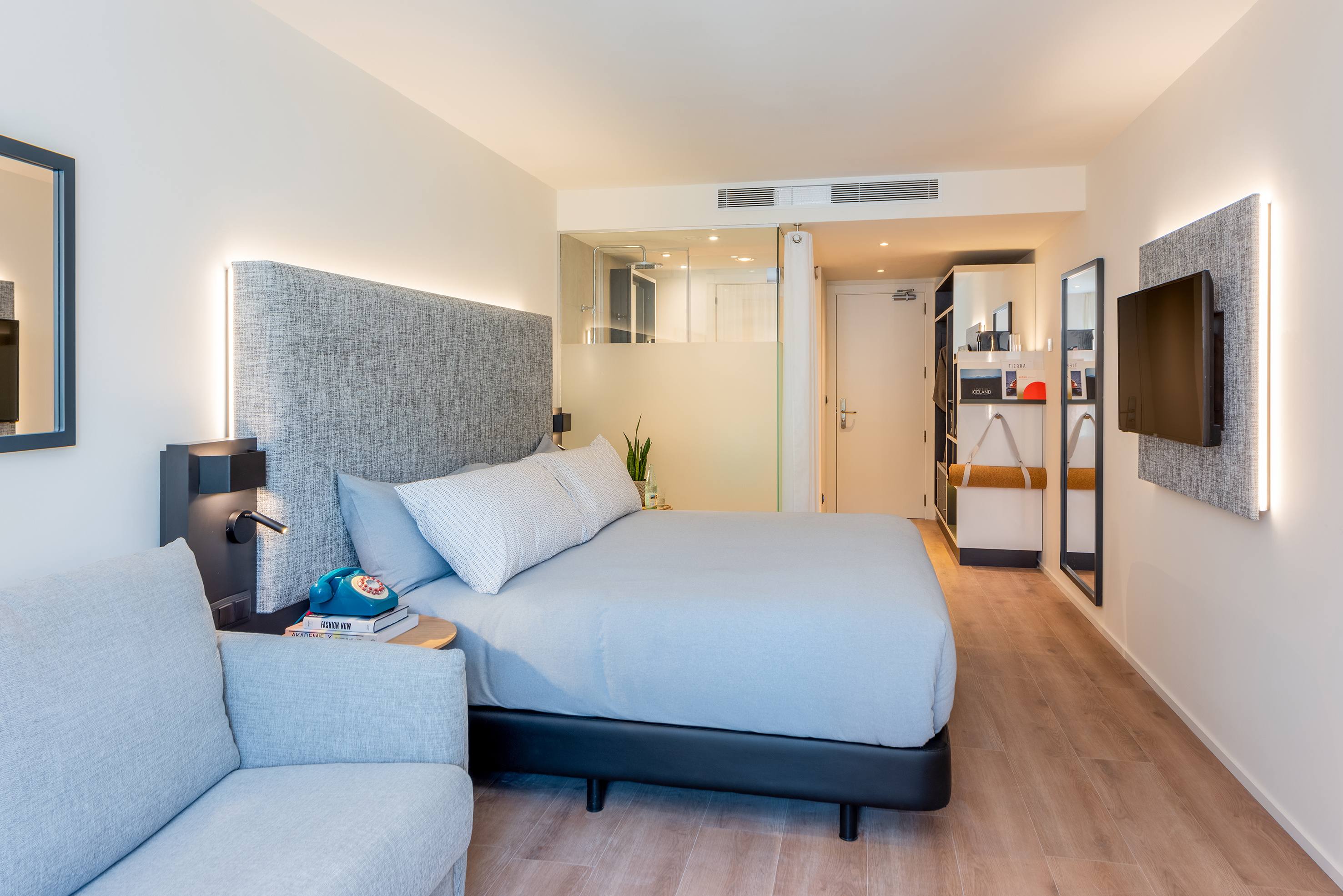 Modern hotel bedroom with king bed, grey headboard, sofa, wall TV, wood floor.