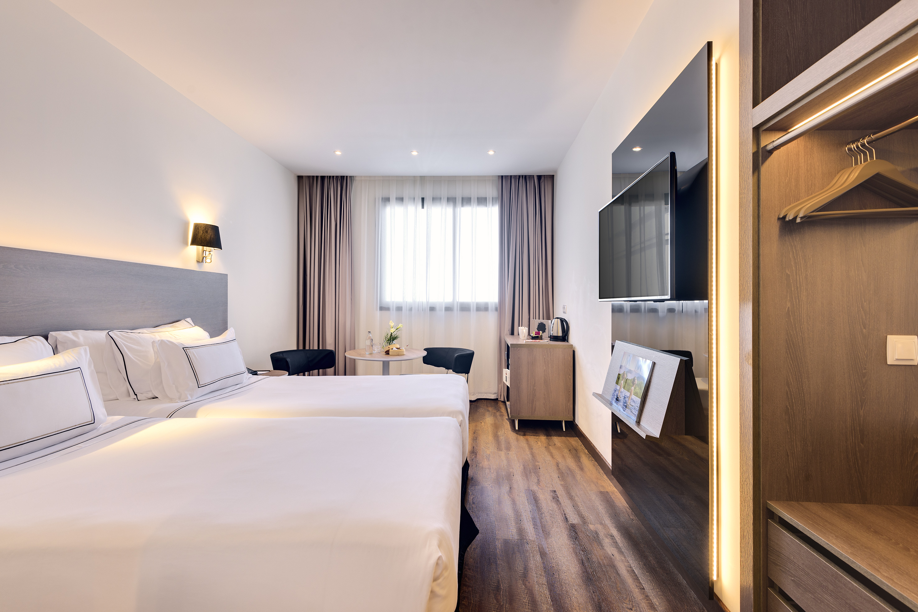Bright modern hotel guest room with two beds, TV, and wardrobe.