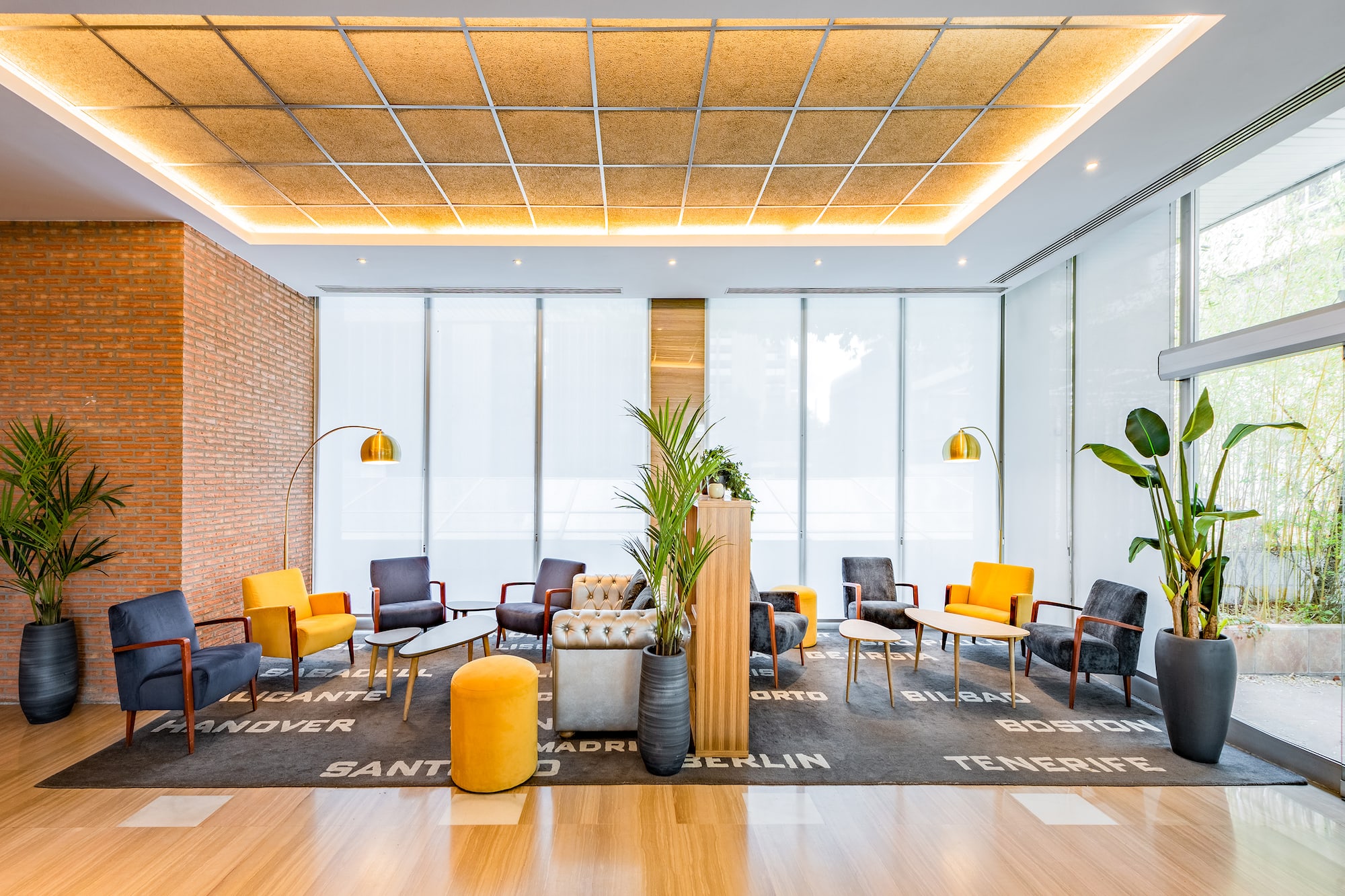 Hotel lobby with modern armchairs, city-name rug, plants, and brick accent wall.
