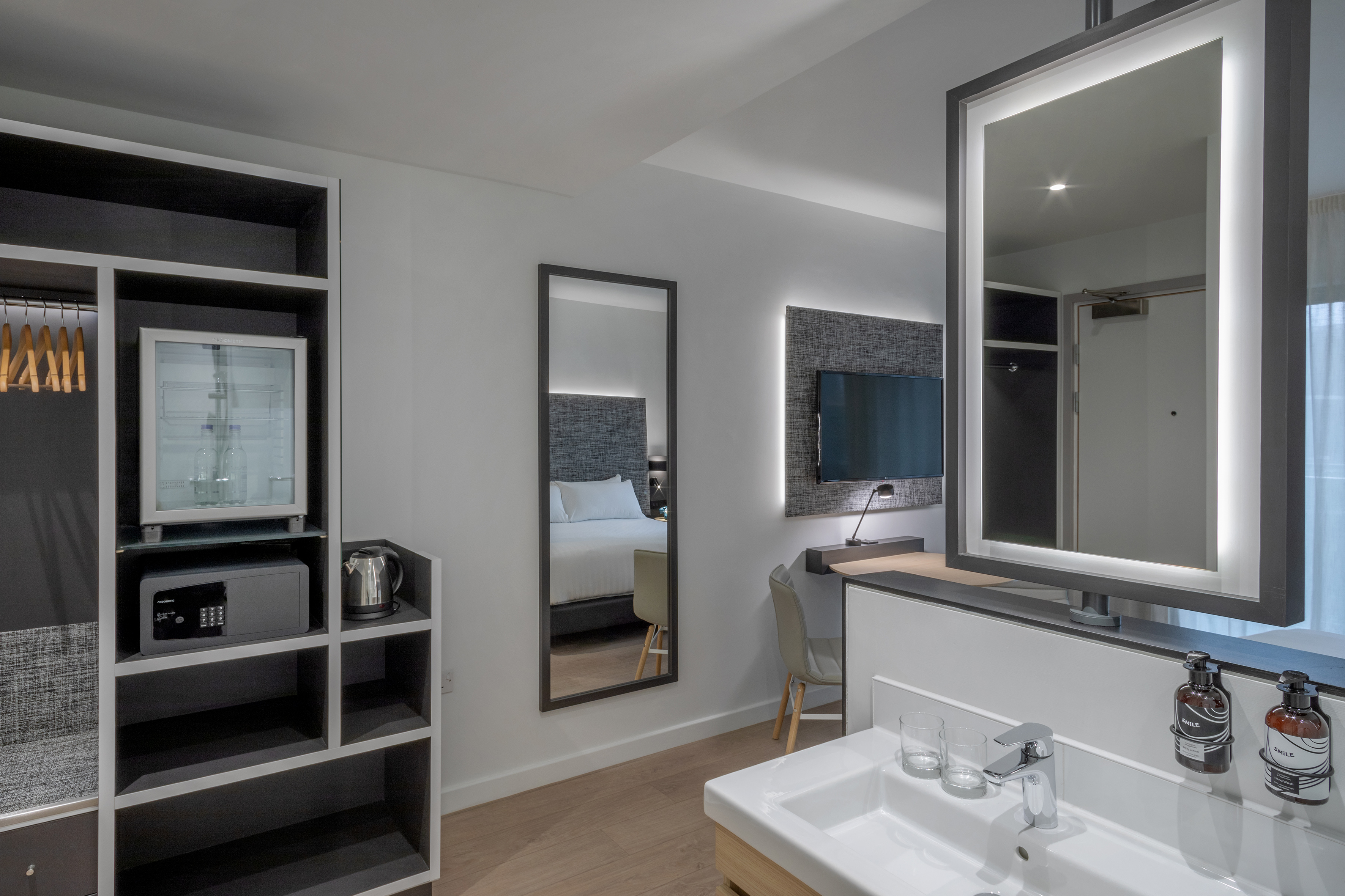 Modern hotel room: vanity, backlit mirror, mini-fridge, safe, and TV.
