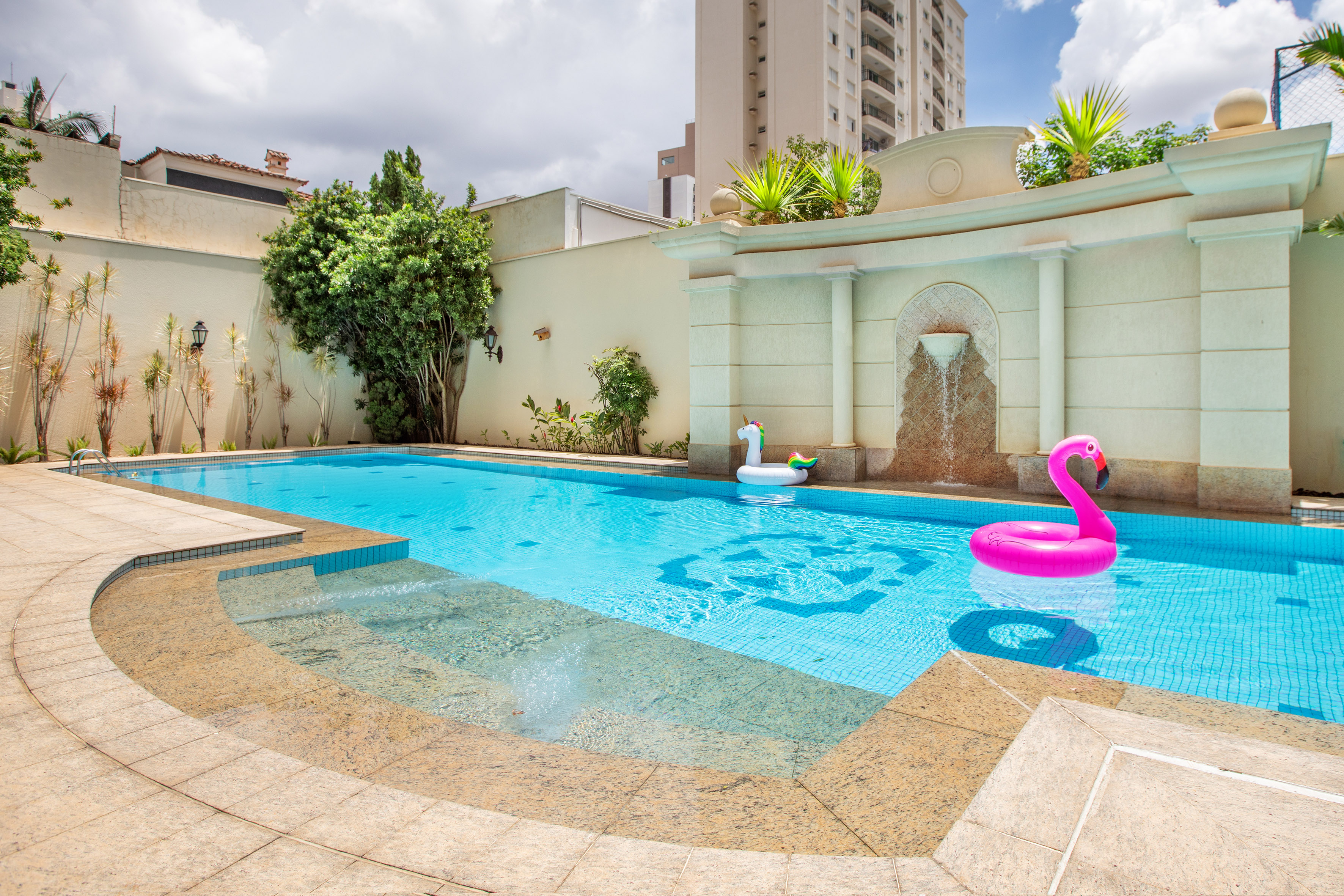 Outdoor pool with flamingo, unicorn floats, waterfall, patio, and building.