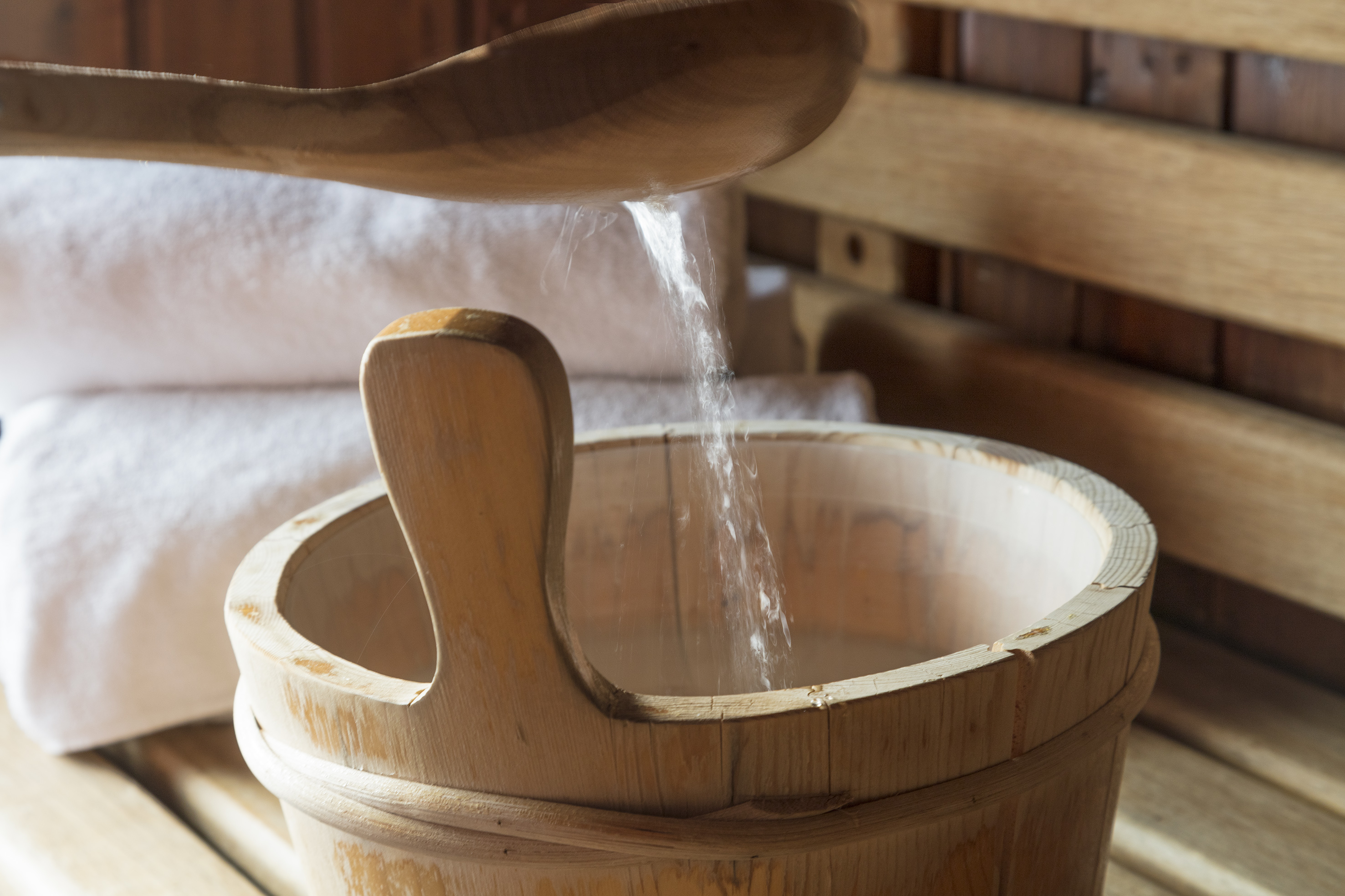 Wooden sauna bucket with water pouring from a ladle, clean towels in background.