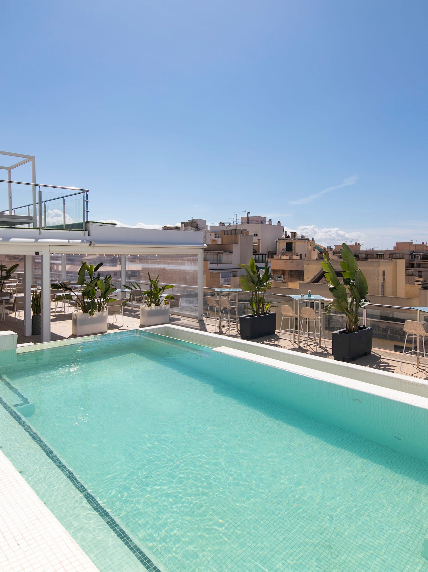Rooftop swimming pool with city view, white stairs, and lounge area under blue sky.