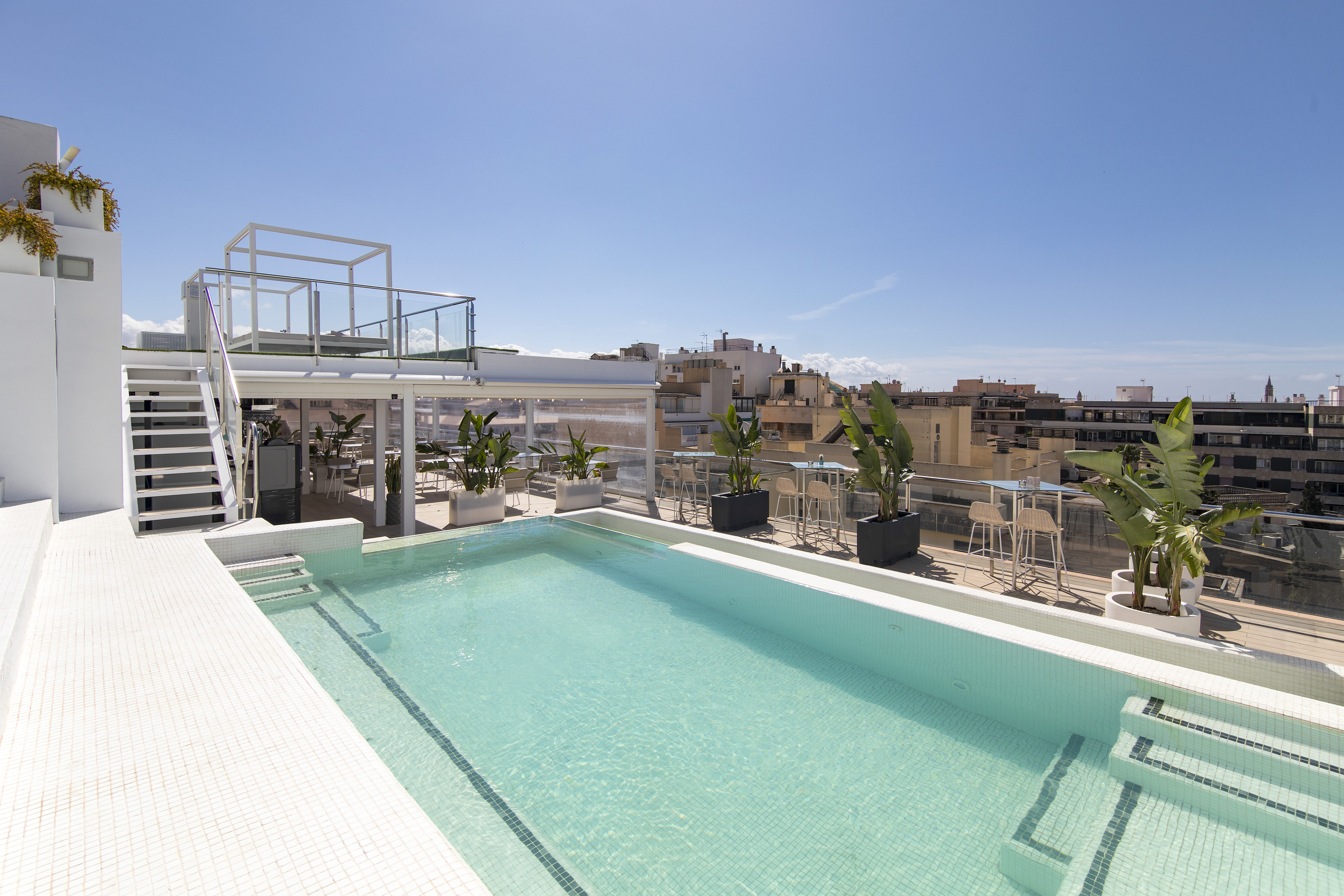 Rooftop swimming pool with city view, white stairs, and lounge area under blue sky.