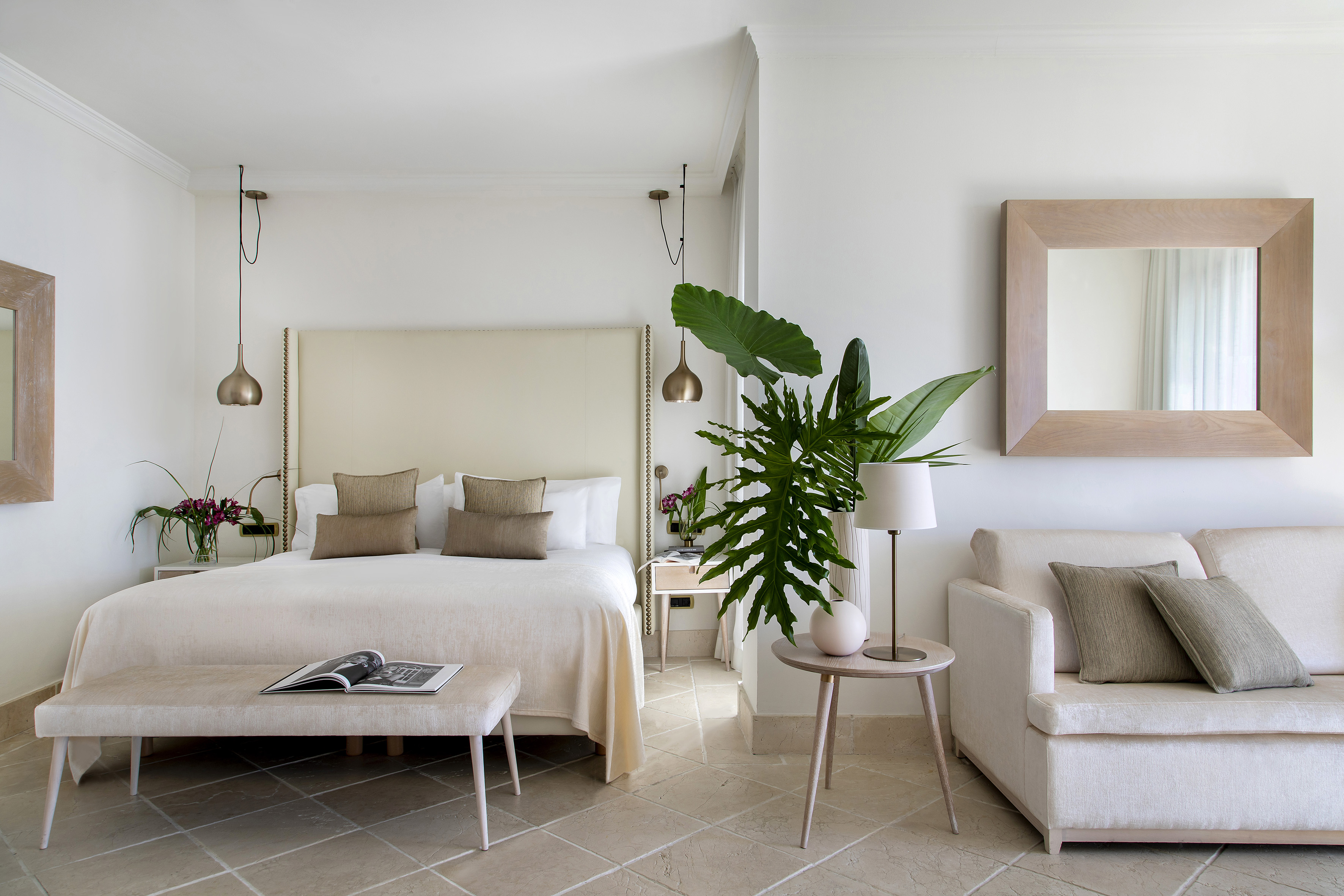 Luxury hotel bedroom with cream bed, sofa, mirror, large plant, and tiled floor.