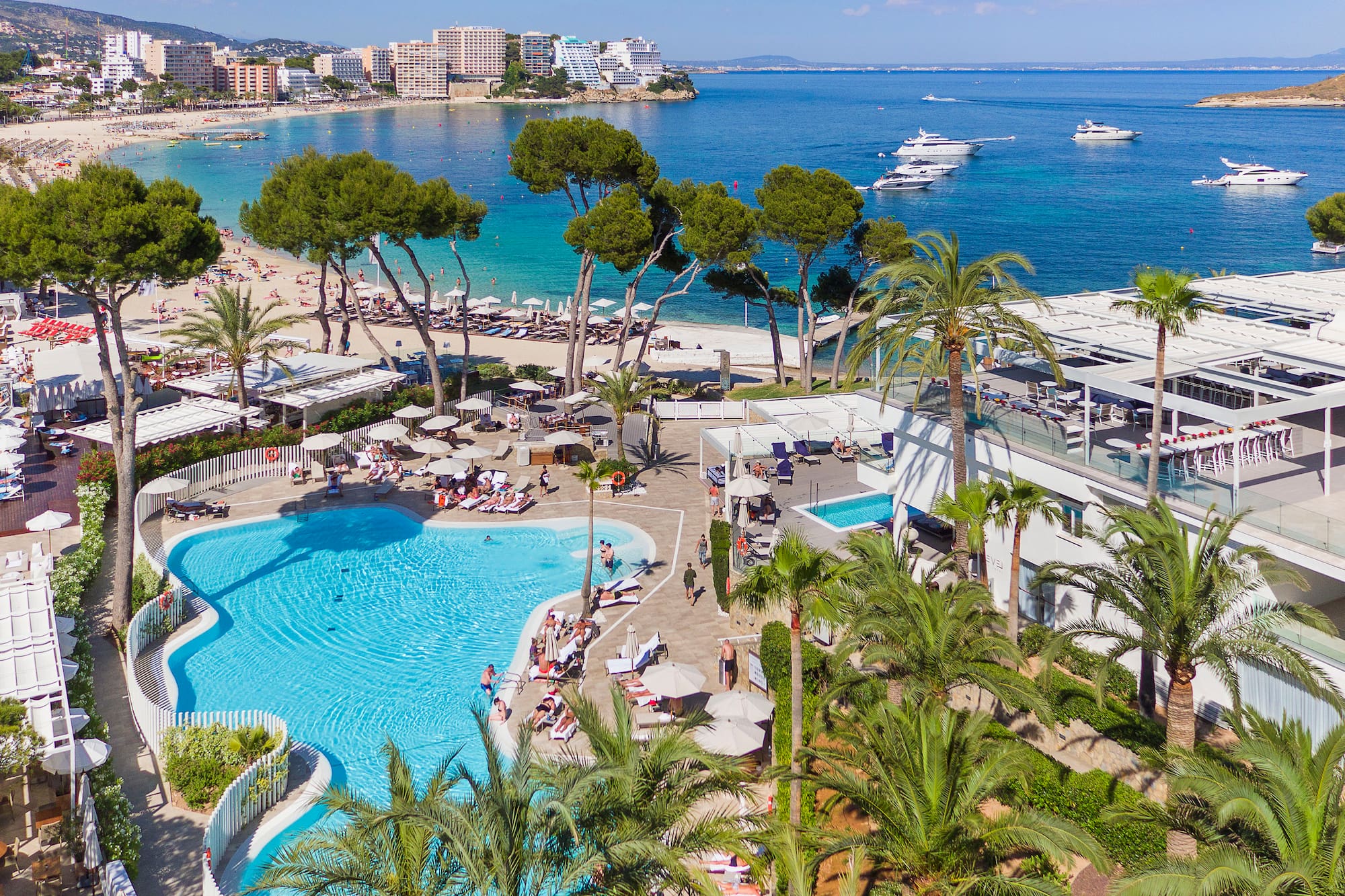 Sunny resort aerial: large pool, beach, palm trees, boats on clear blue ocean.