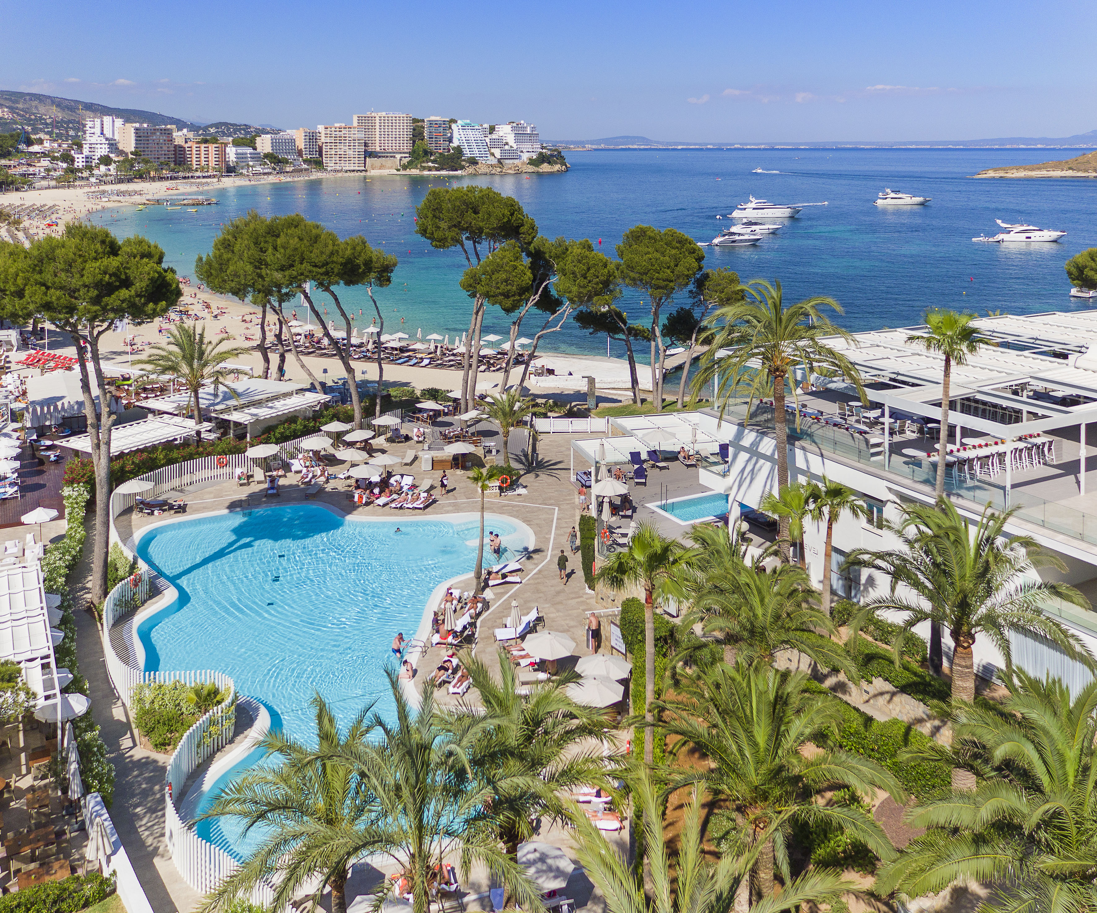 Sunny resort aerial: large pool, beach, palm trees, boats on clear blue ocean.