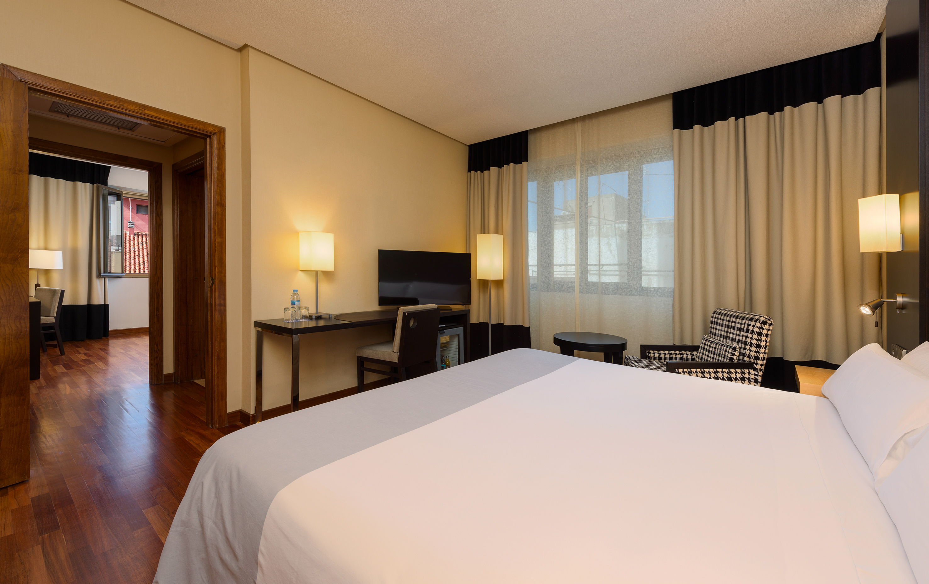 Modern hotel suite bedroom with large bed, desk, TV, and sitting area.