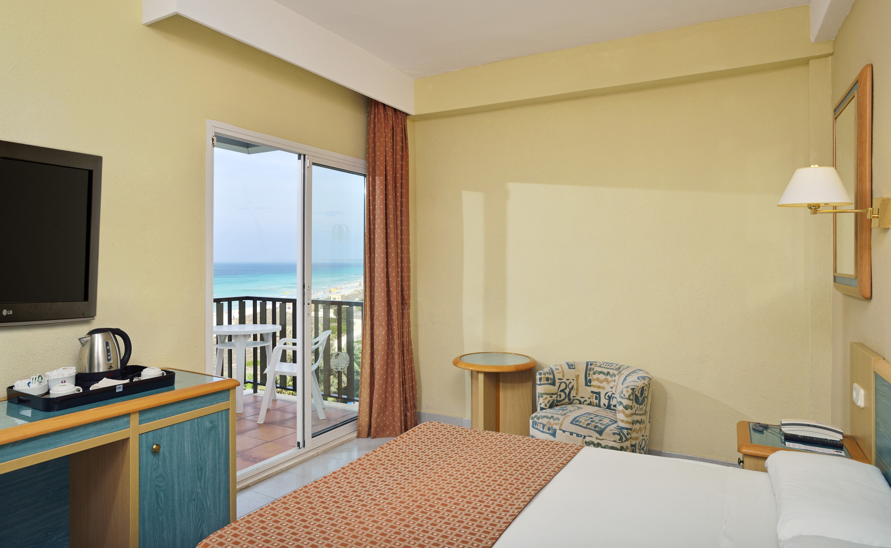 Hotel room with ocean view balcony, beach, bed, and TV.
